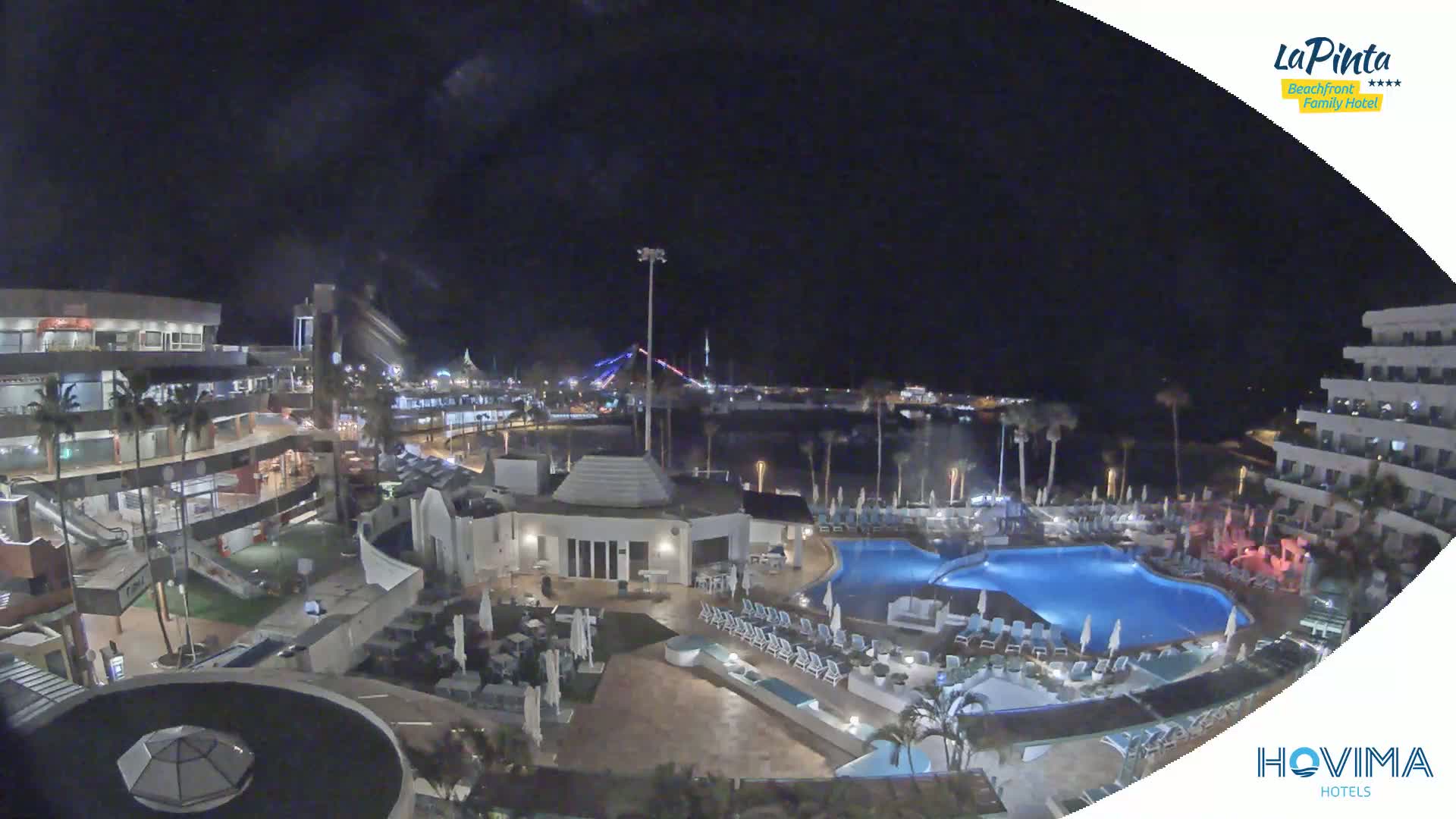 A nighttime aerial view of a resort complex with swimming pools, palm trees, and buildings, overlooking a calm body of water under a clear, dark sky.
