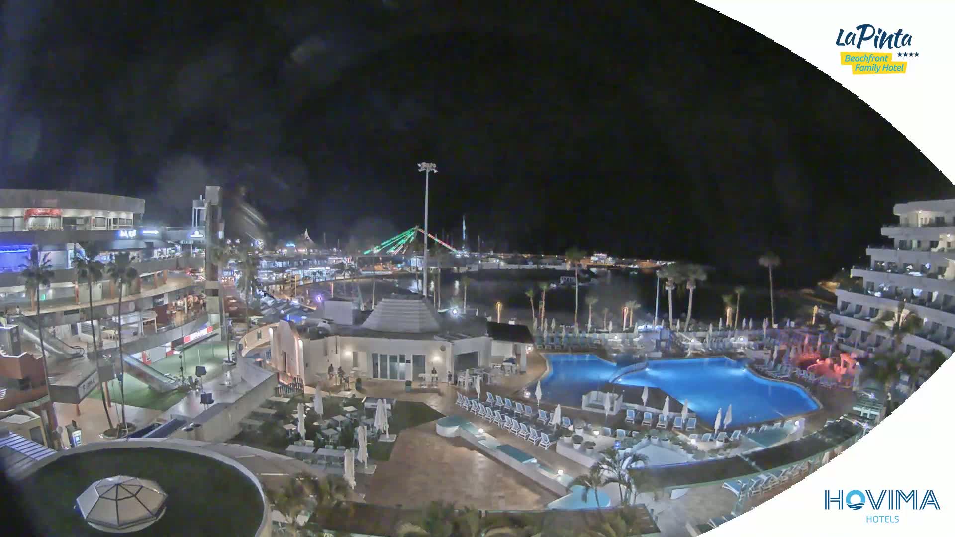 A nighttime aerial view of a resort complex with illuminated pools and surrounding buildings, overlooking a harbor with a bridge and palm trees under a dark, clear sky.