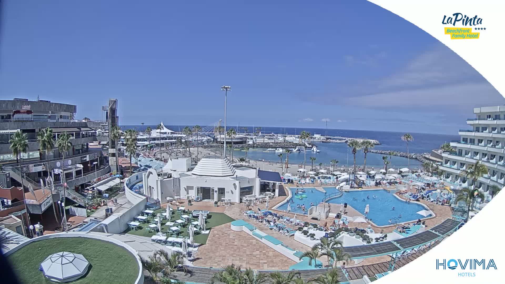 A sunny coastal resort scene shows a large swimming pool, beach, marina, and several buildings, with people relaxing and swimming.