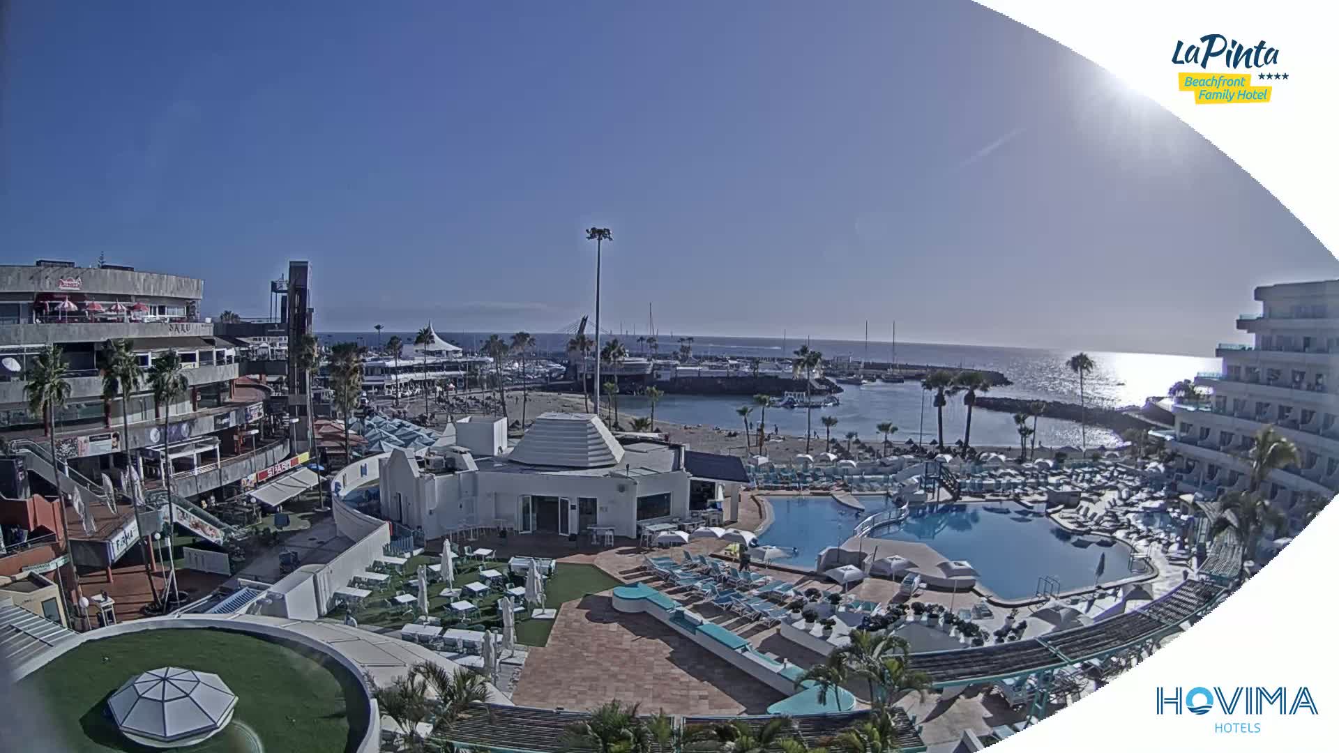 A sunny day overlooks a resort with a swimming pool, beach, and marina, situated near buildings and palm trees.