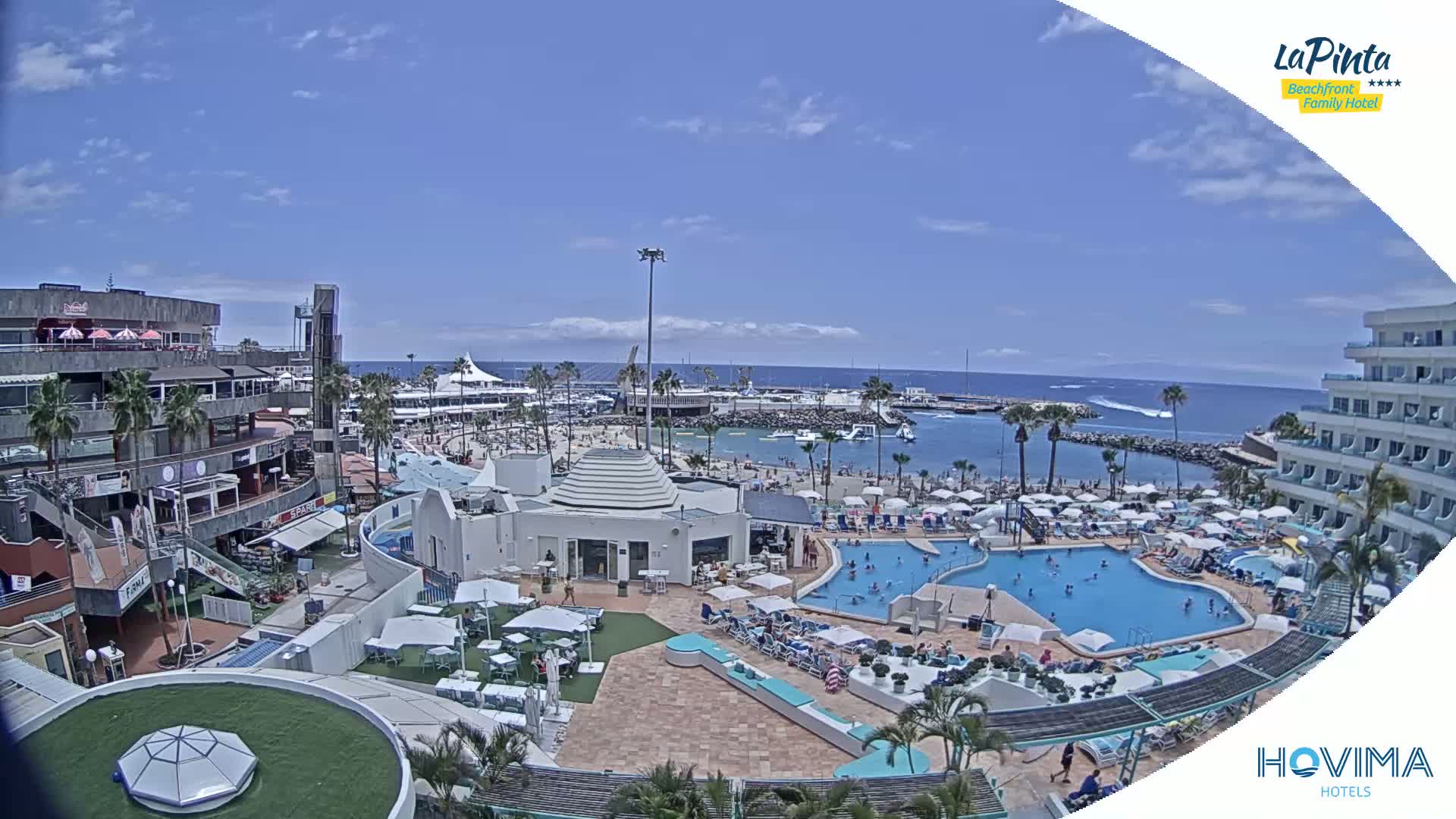 A resort with multiple pools is shown on a sunny day next to a calm ocean; a beach and marina are visible in the background.