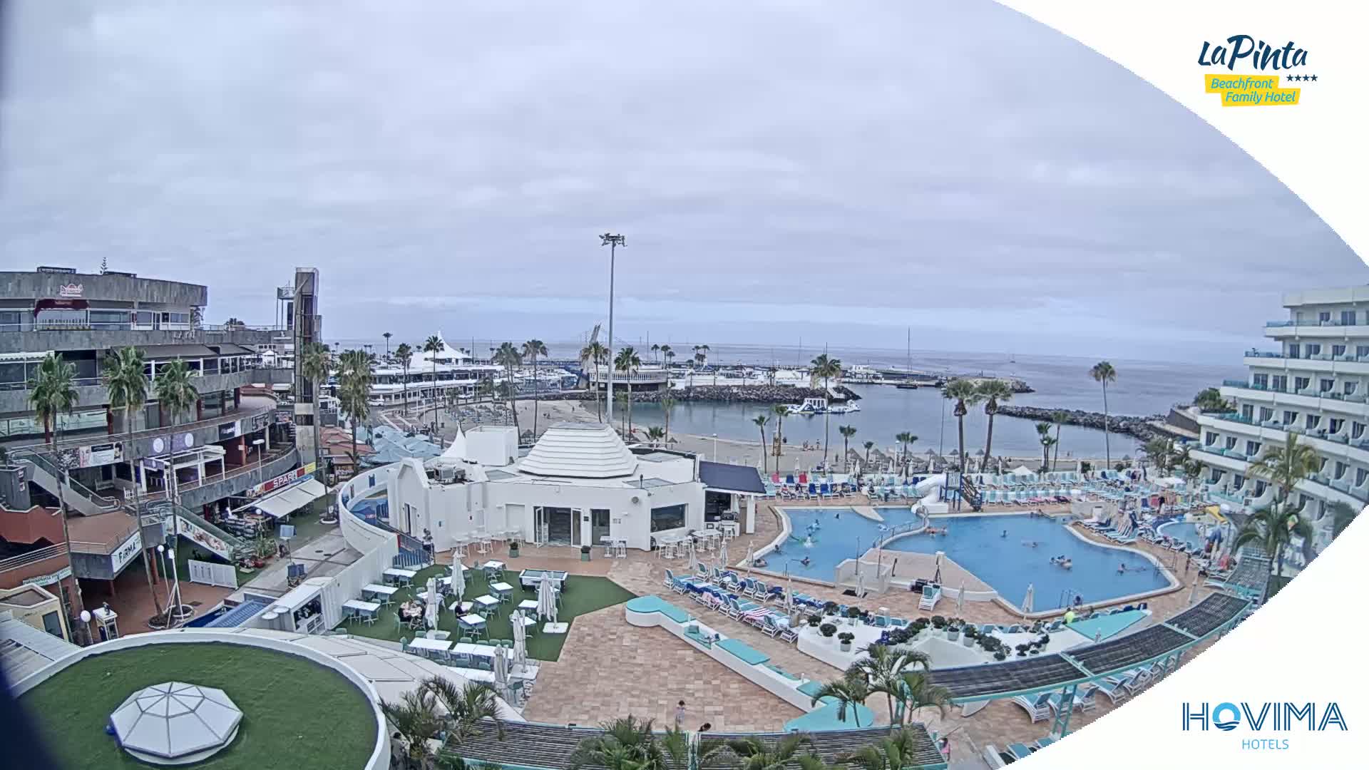 An overcast day reveals a resort with a swimming pool and surrounding buildings near a marina and ocean.
