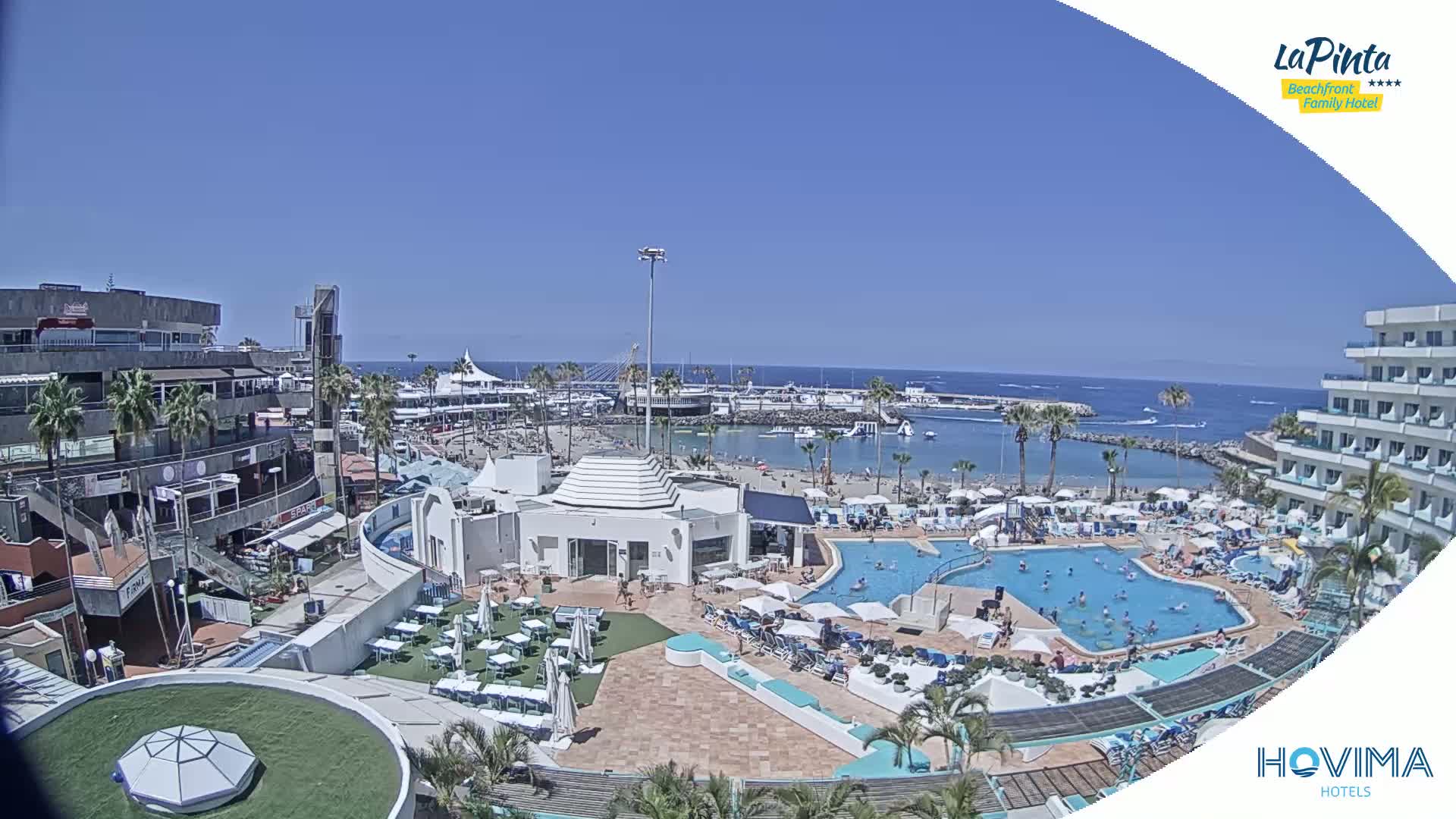 A sunny day overlooking a resort with multiple pools, a beach, and a marina, all situated near the ocean.