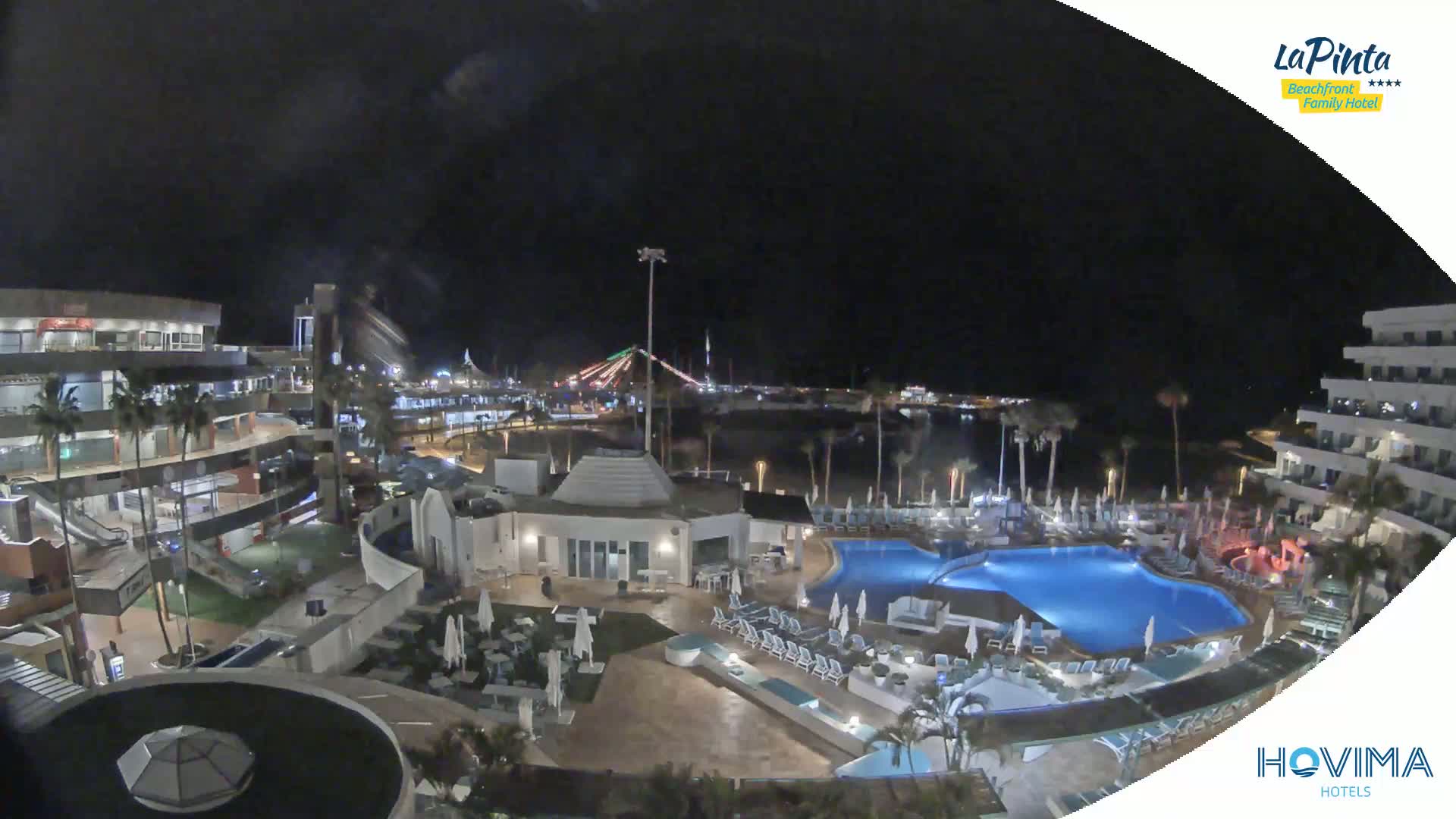 A nighttime aerial view of a resort complex with a swimming pool, surrounding buildings, and palm trees, overlooking a dark body of water under a clear sky.