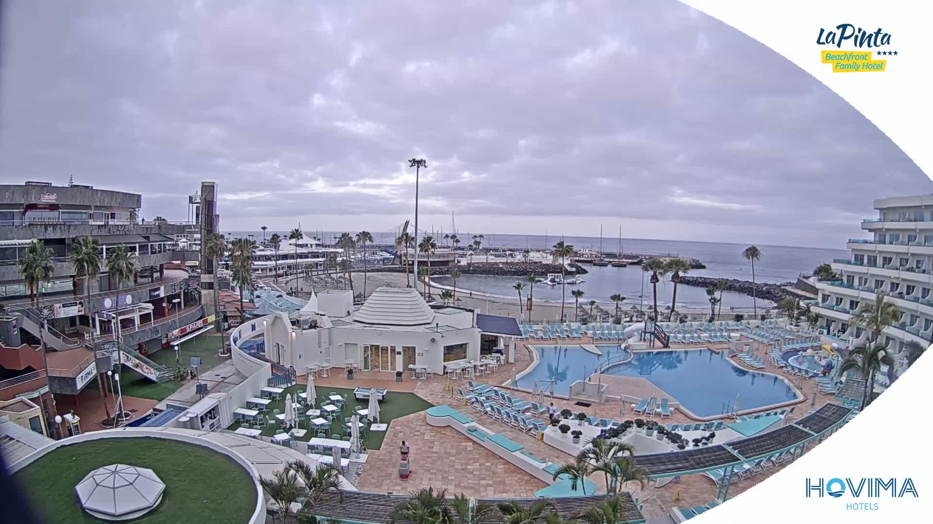 An overcast day reveals a resort with multiple pools overlooking a calm bay and a small beach, adjacent to shops and buildings.