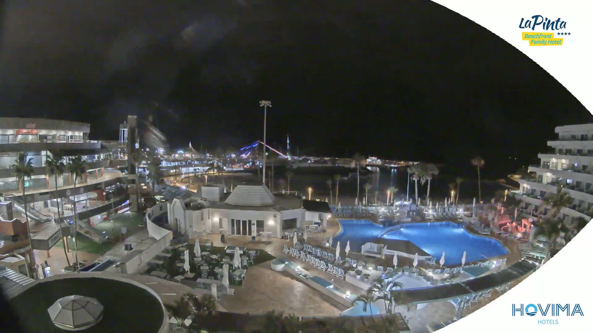 A nighttime aerial view of a resort complex with a swimming pool, situated near a waterfront with palm trees under a clear, dark sky.