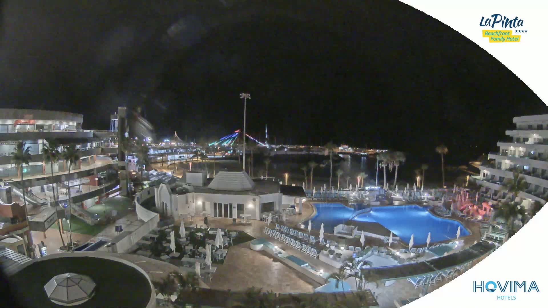 A nighttime view of a resort area with a large pool and palm trees, overlooking a calm body of water and a brightly lit waterfront area under a dark sky.