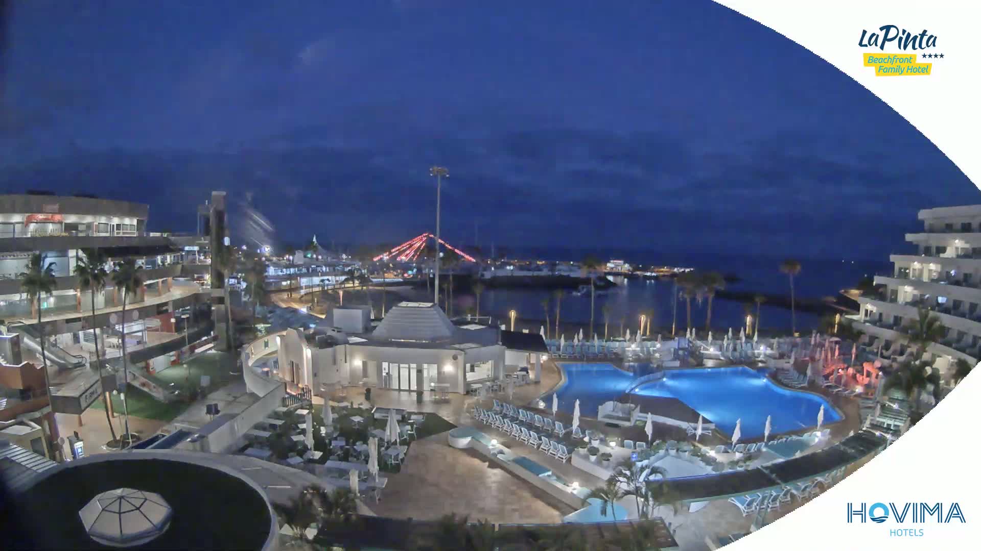 An illuminated resort complex at night overlooks a calm body of water under a dark blue, mostly clear sky.