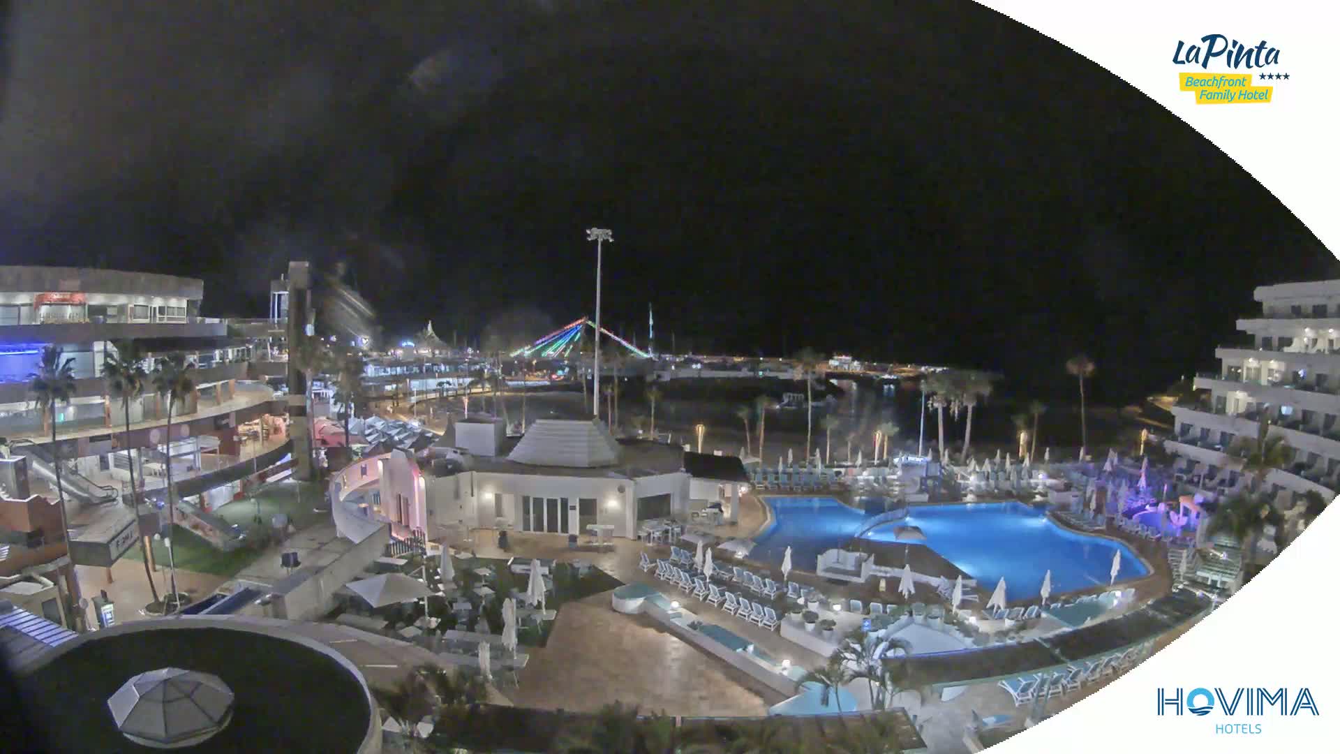 A nighttime view of a resort complex with swimming pools, surrounding buildings, and a brightly lit bridge in the distance.