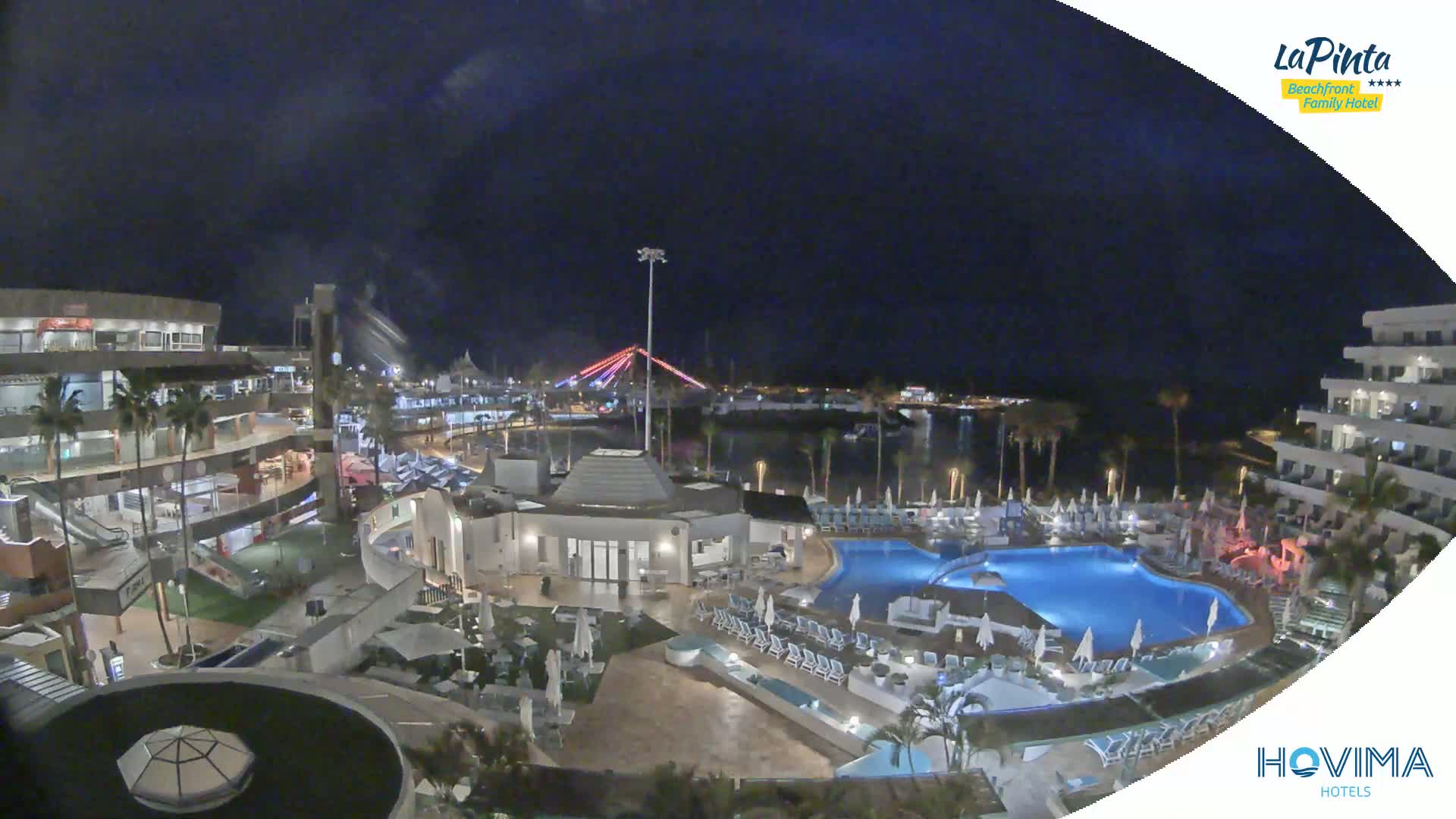 A nighttime aerial view of a resort complex with pools and surrounding buildings, under a dark, cloudy sky.