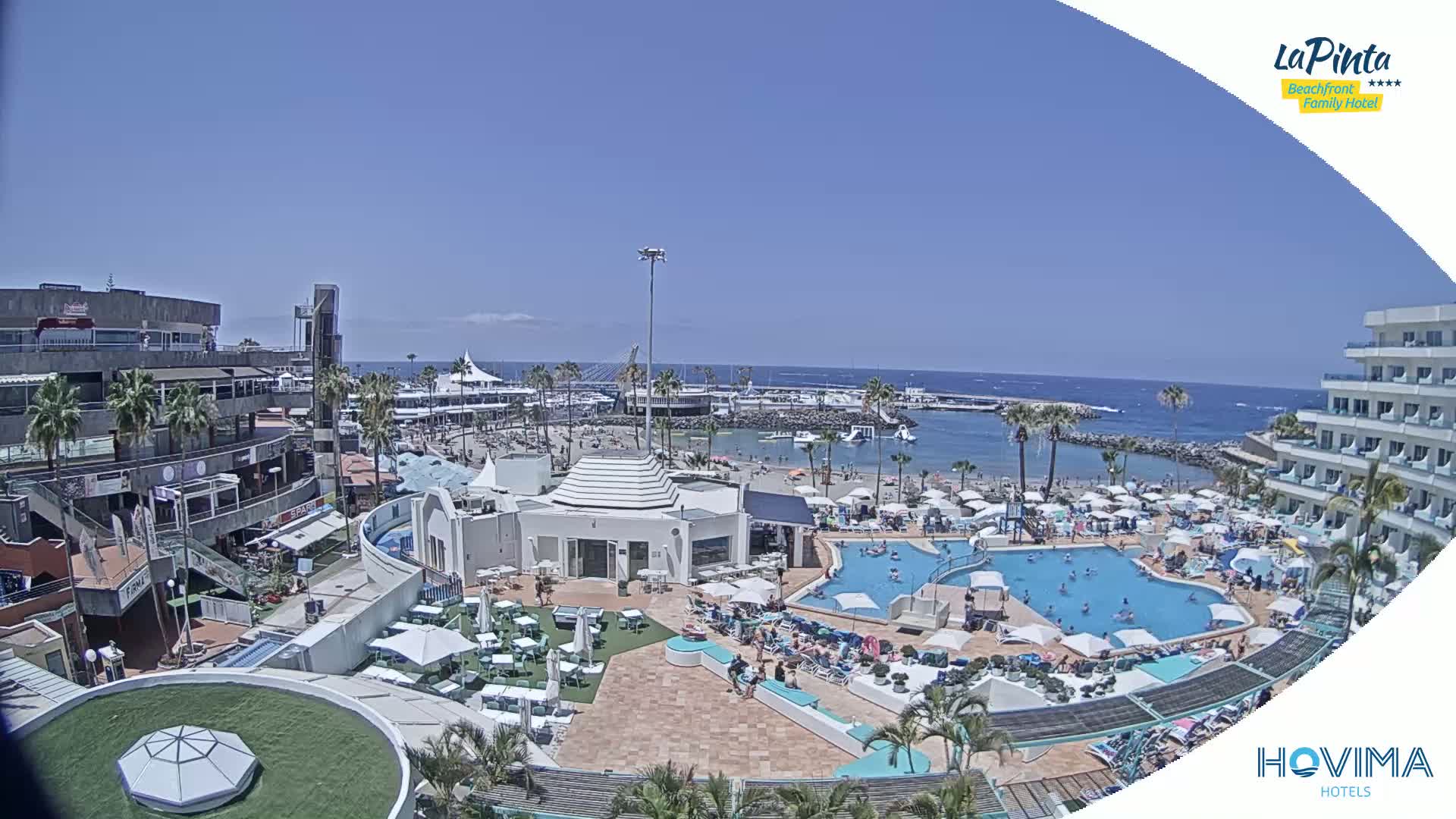 An aerial view of a resort complex on a sunny day, showing a swimming pool, beach area, and buildings overlooking a calm ocean.
