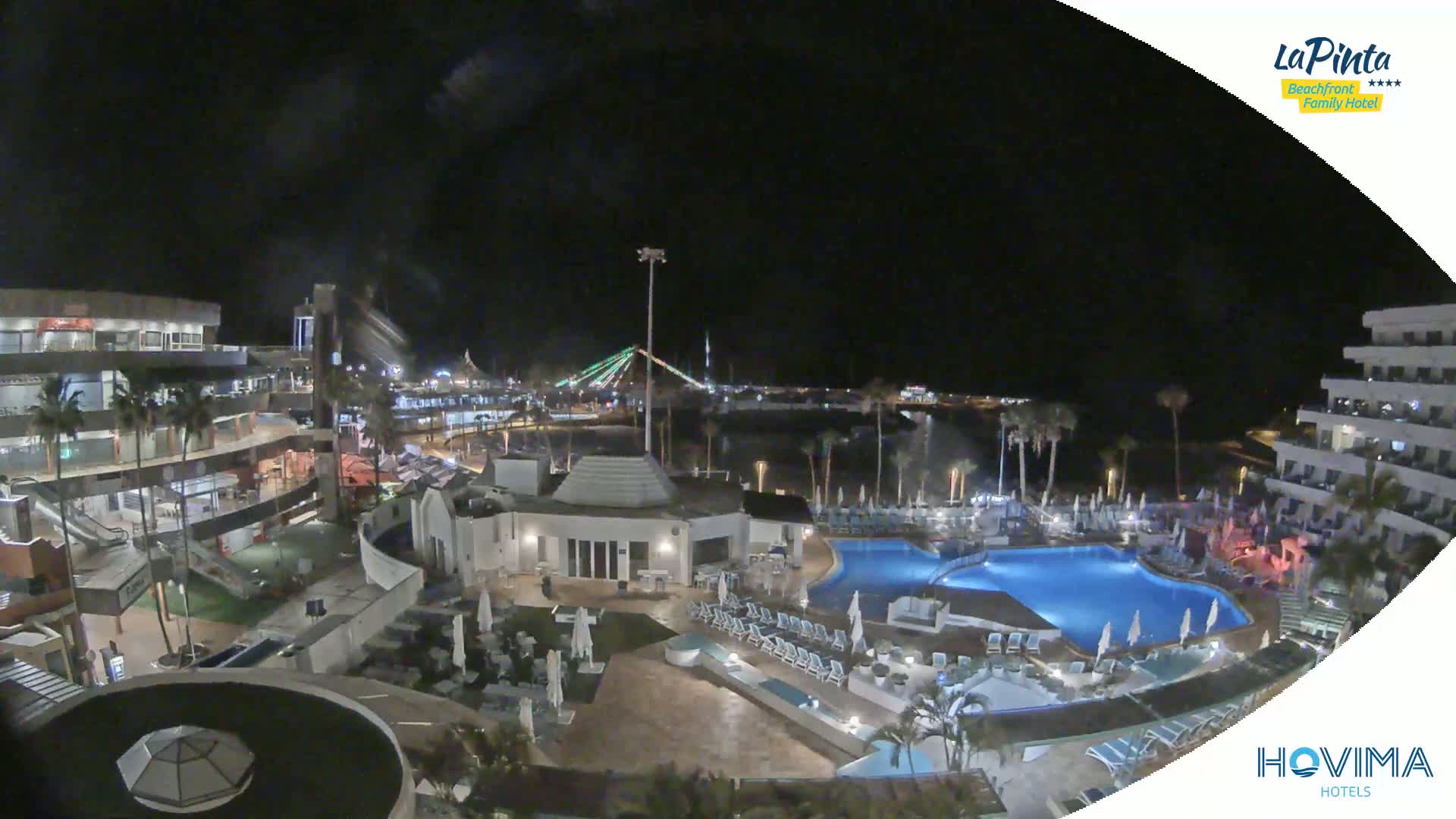 A nighttime aerial view of a resort complex with swimming pools, surrounding buildings, and a bridge in the distance under a clear, dark sky.