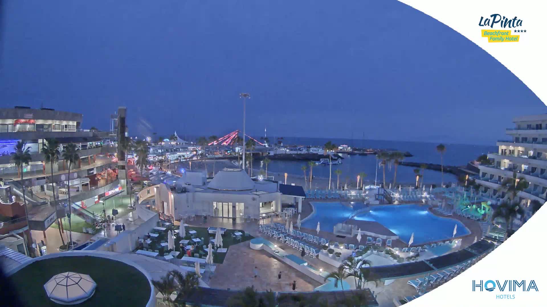 A nighttime aerial view of a resort complex with pools and a beachfront area, under a twilight sky.