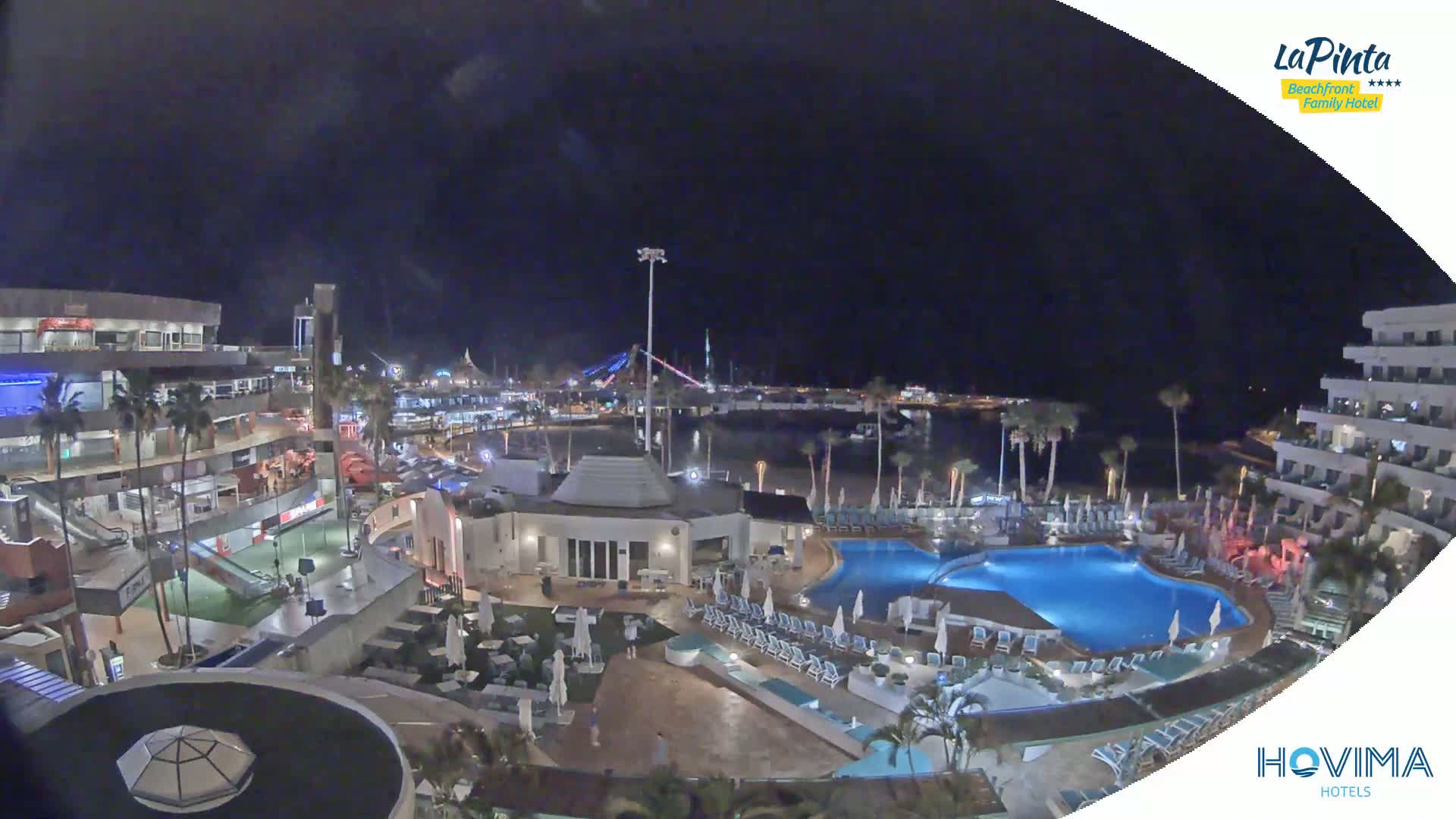 A nighttime aerial view of a resort complex with pools and surrounding buildings, overlooking a calm harbor under a dark sky.