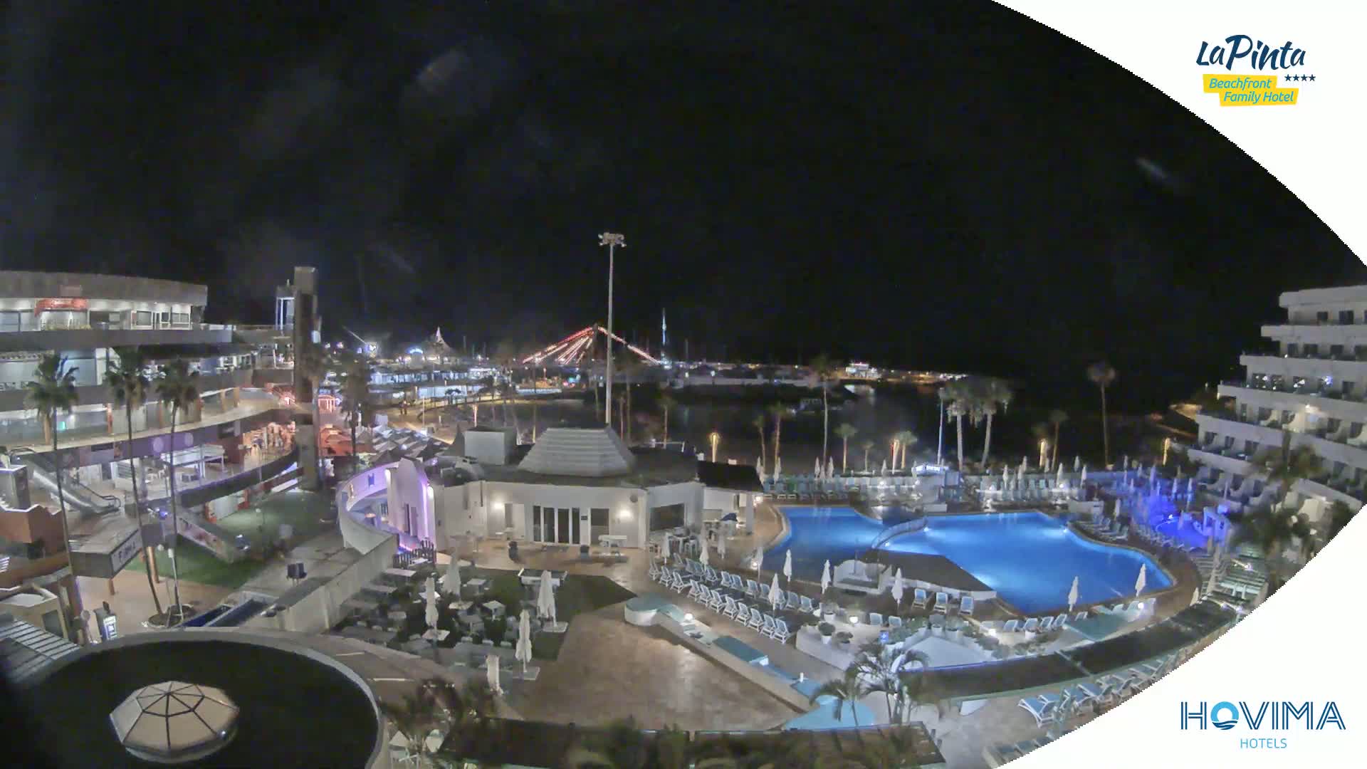 A nighttime aerial view of a resort complex with swimming pools, surrounding buildings, and palm trees, overlooking a calm body of water and a city skyline under a clear, dark sky.