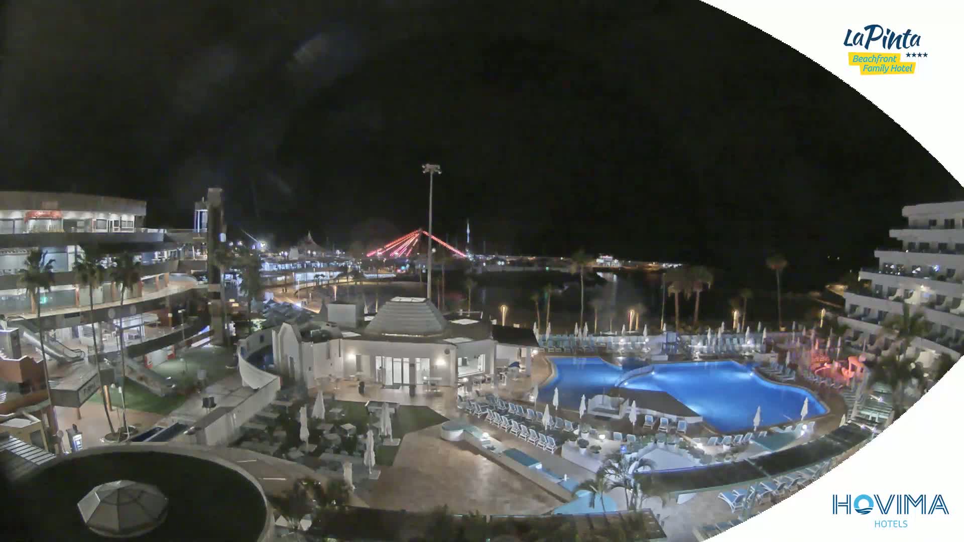 A nighttime aerial view of a resort complex with swimming pools, lit buildings, and palm trees, overlooking a body of water with a bridge in the distance.