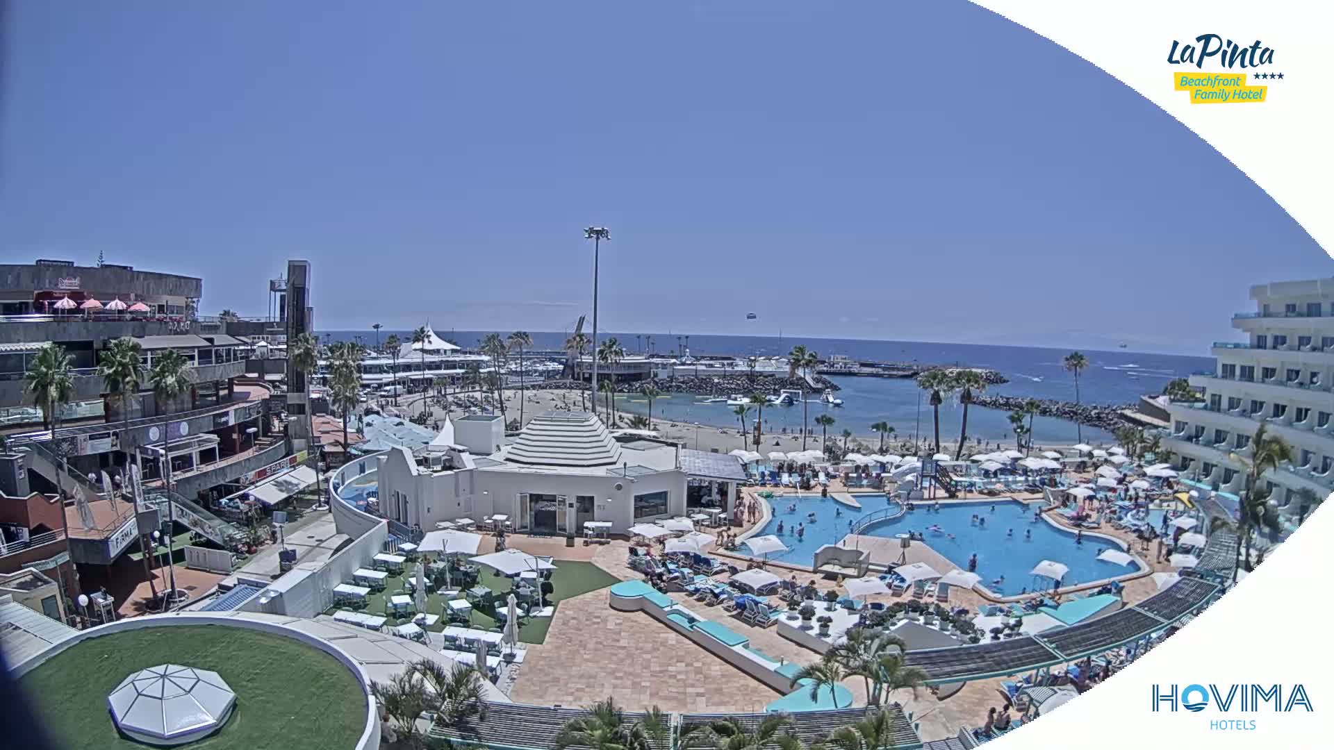 A resort with a large pool and beach area is shown on a sunny day, overlooking a calm ocean.
