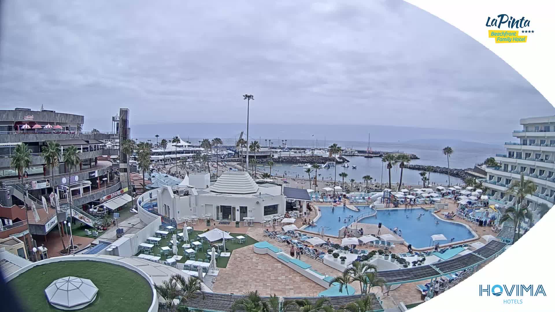 An overcast day shows a resort area with a swimming pool, beach, marina, and shops, all situated next to the ocean.