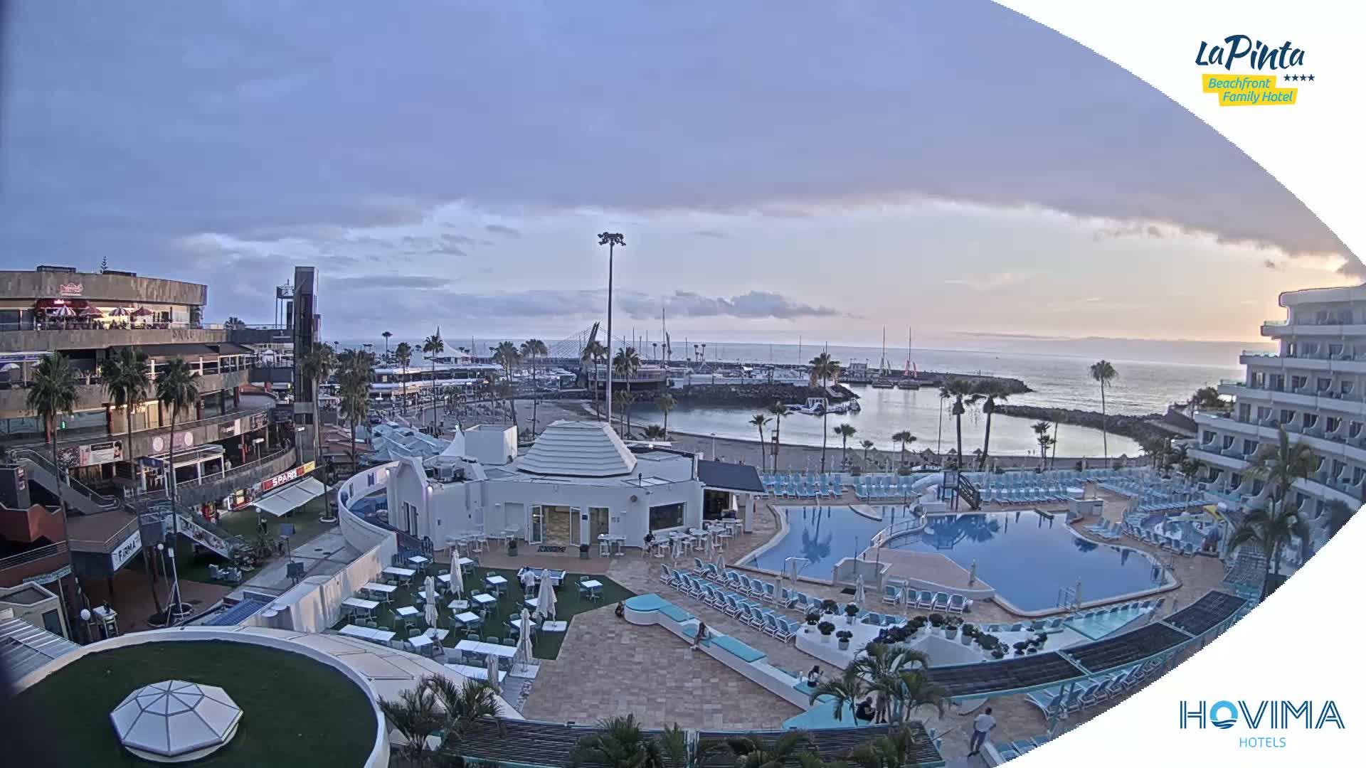 An overcast evening scene shows a resort area with multiple pools, a beach, a marina with boats, and buildings overlooking the ocean.