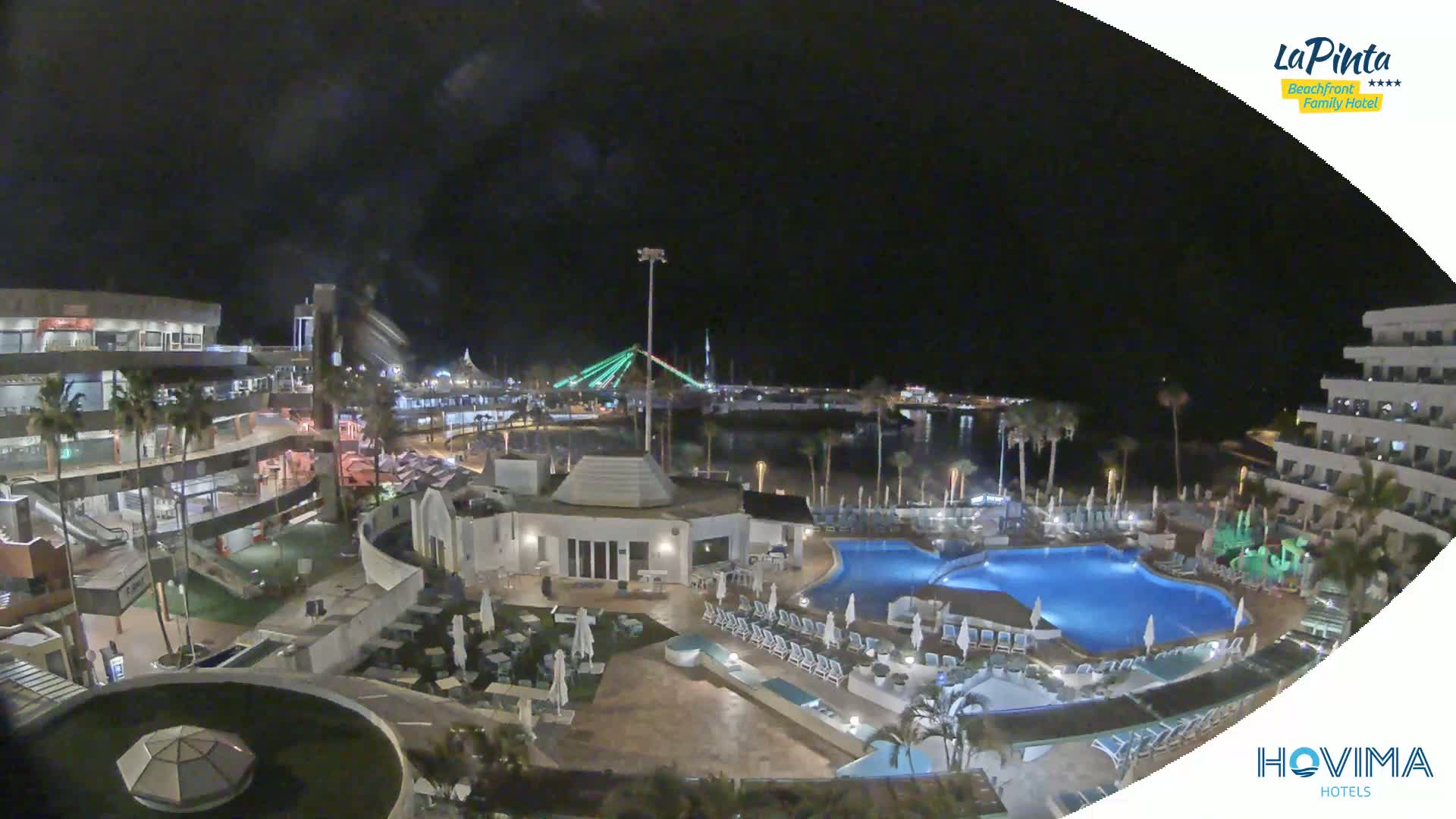 A nighttime aerial view of a resort complex with swimming pools, illuminated buildings, and a waterfront with a bridge in the background under a dark, clear sky.