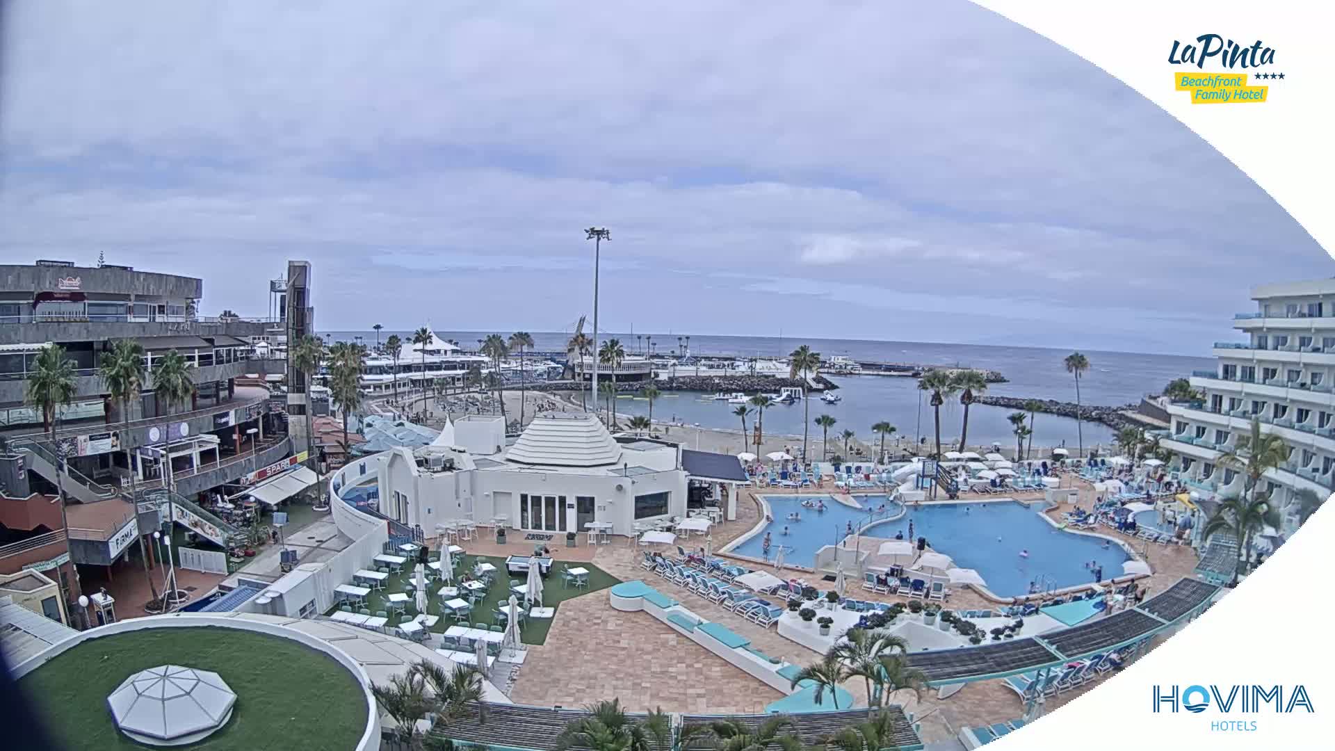 An aerial view shows a resort with multiple pools, a beach, a marina, and shops under a mostly cloudy sky.