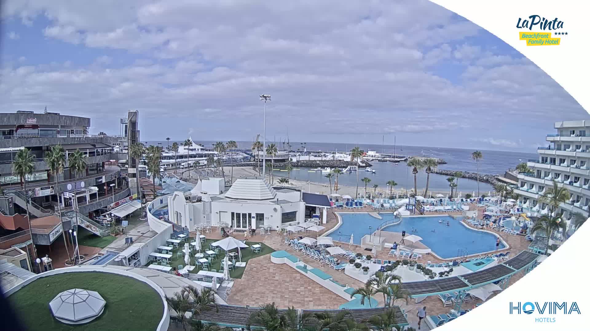 An outdoor scene on a partly cloudy day shows a resort with a swimming pool, surrounding buildings, a marina, and ocean in the background.
