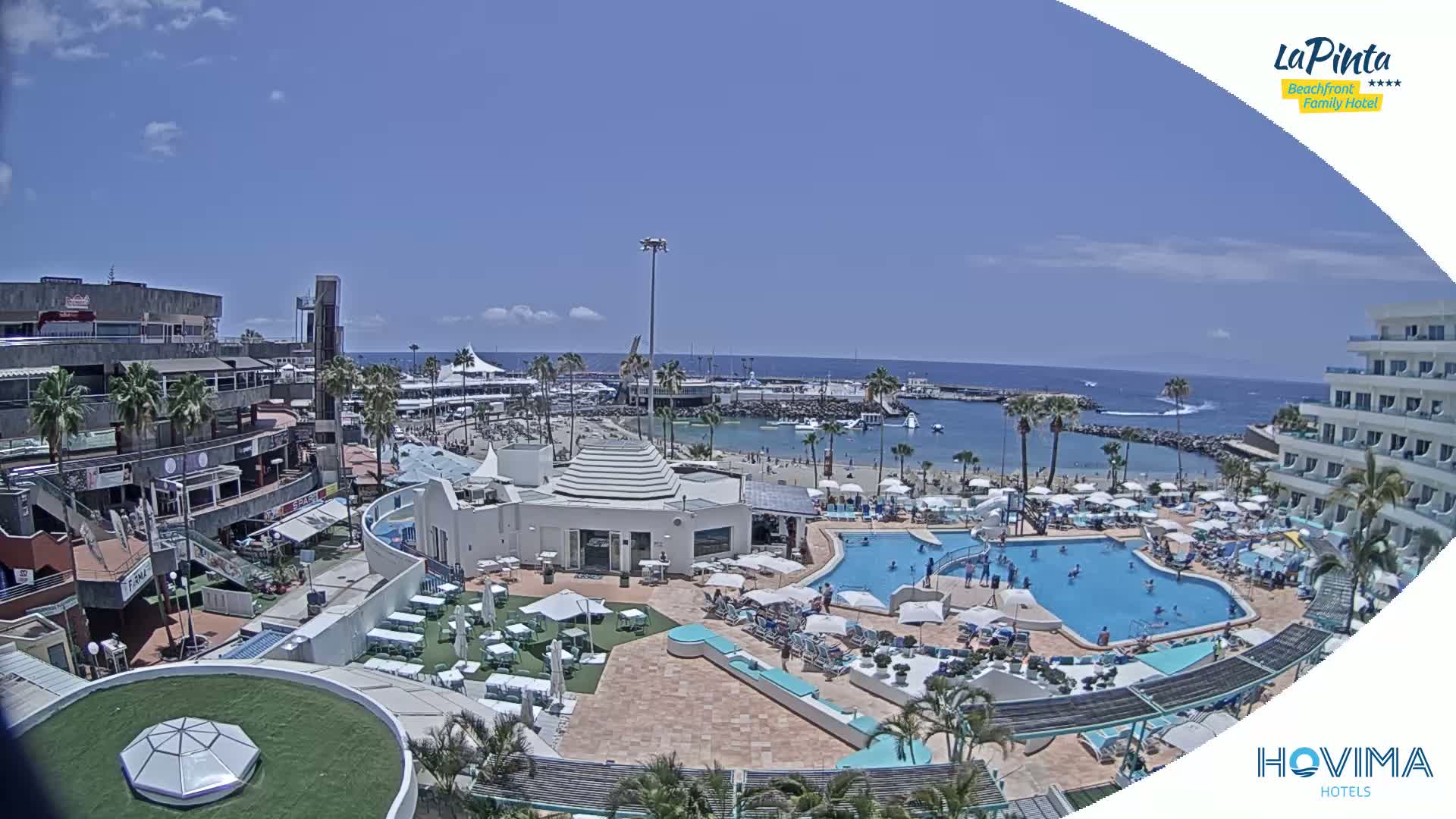 A sunny day overlooking a resort with multiple pools, a beach, and a harbor, all situated near buildings and shops.