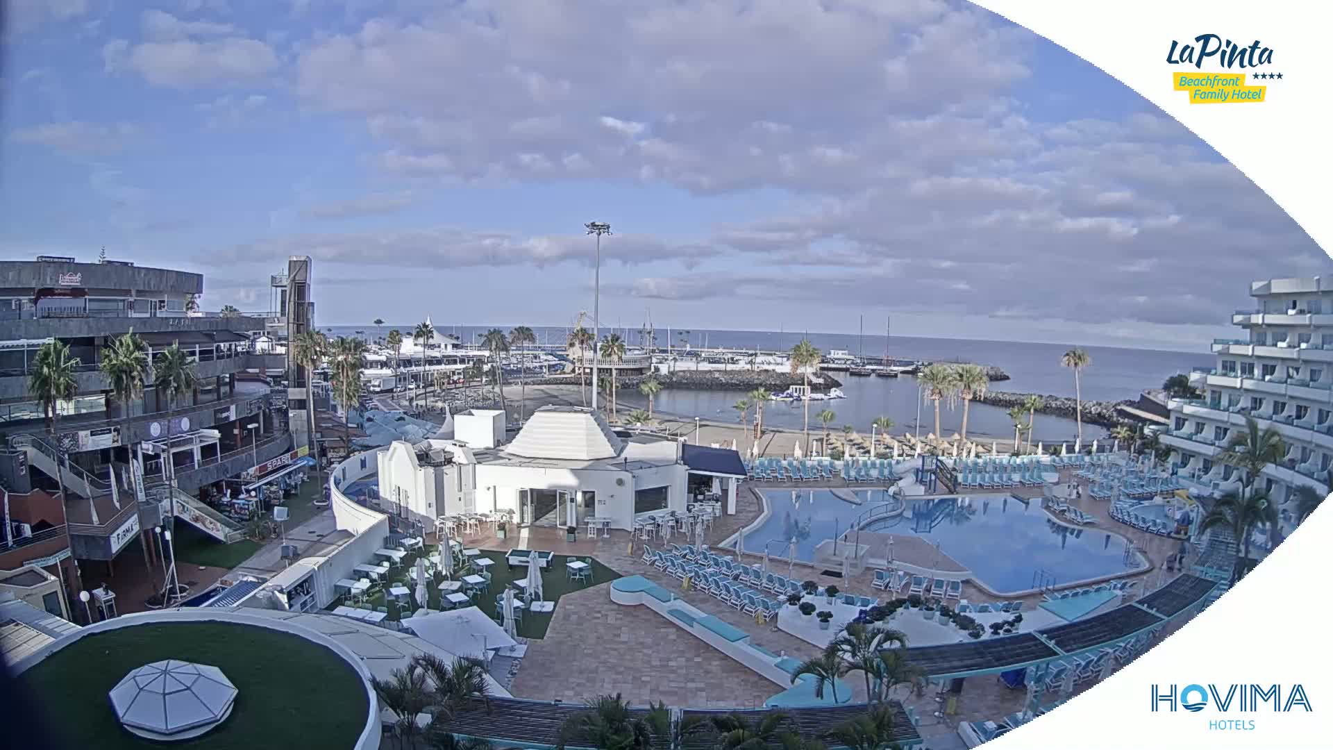 A resort with a swimming pool overlooks a harbor filled with boats under a partly cloudy sky.