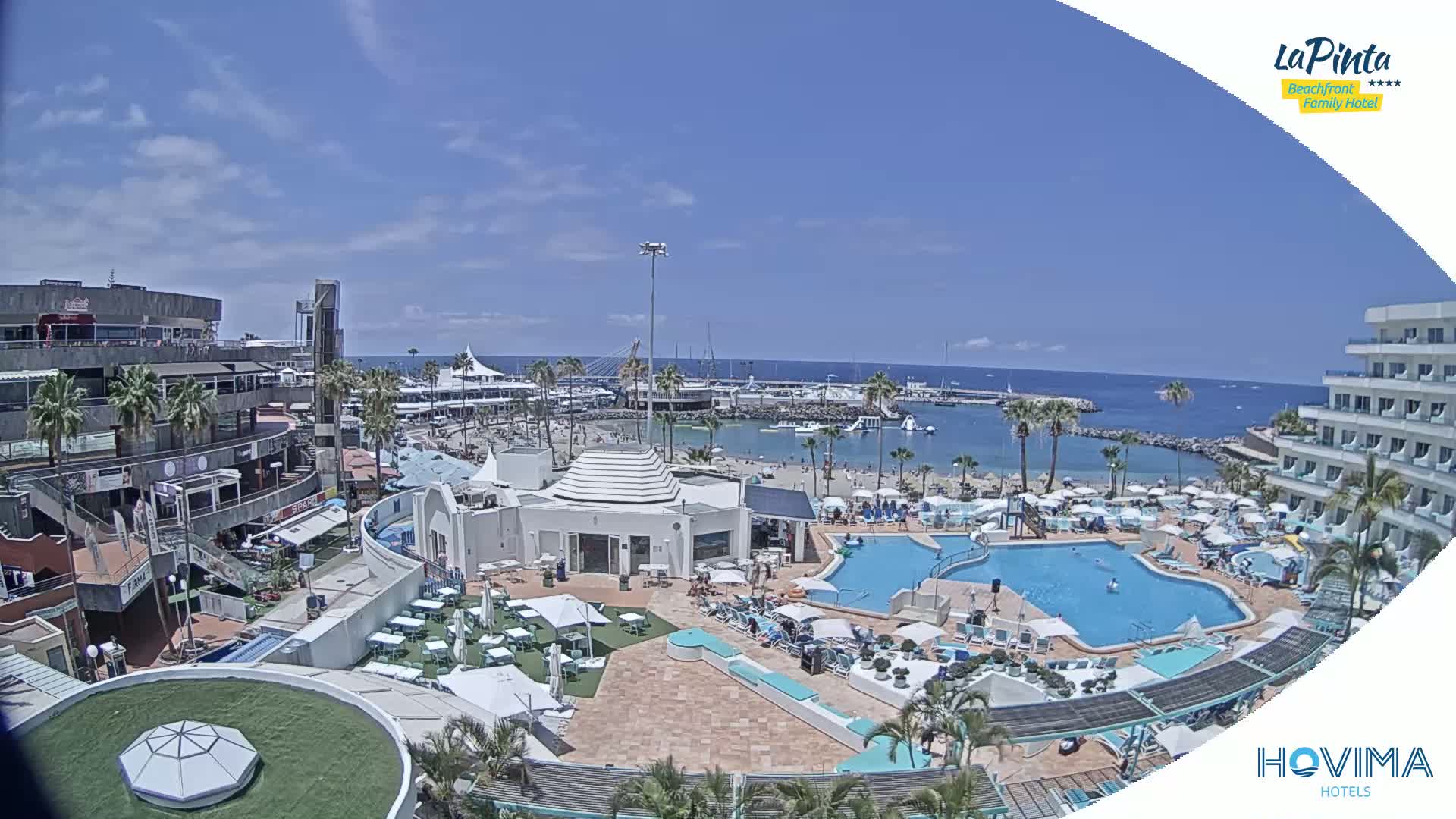 An aerial view shows a sunny resort area with a swimming pool, beach, marina, and buildings, overlooking a calm ocean.