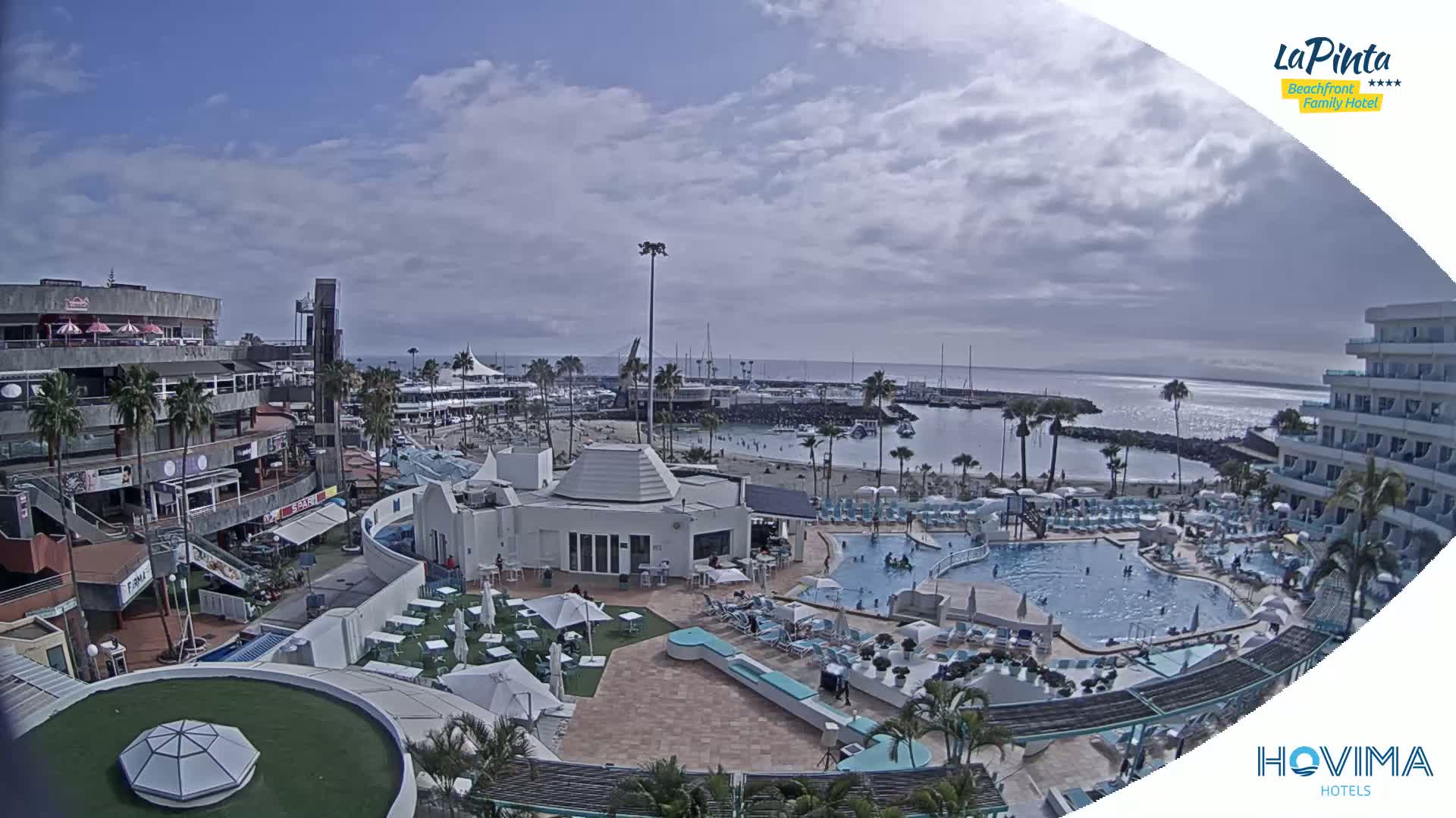 A resort area with pools, a beach, and buildings under a partly cloudy sky.