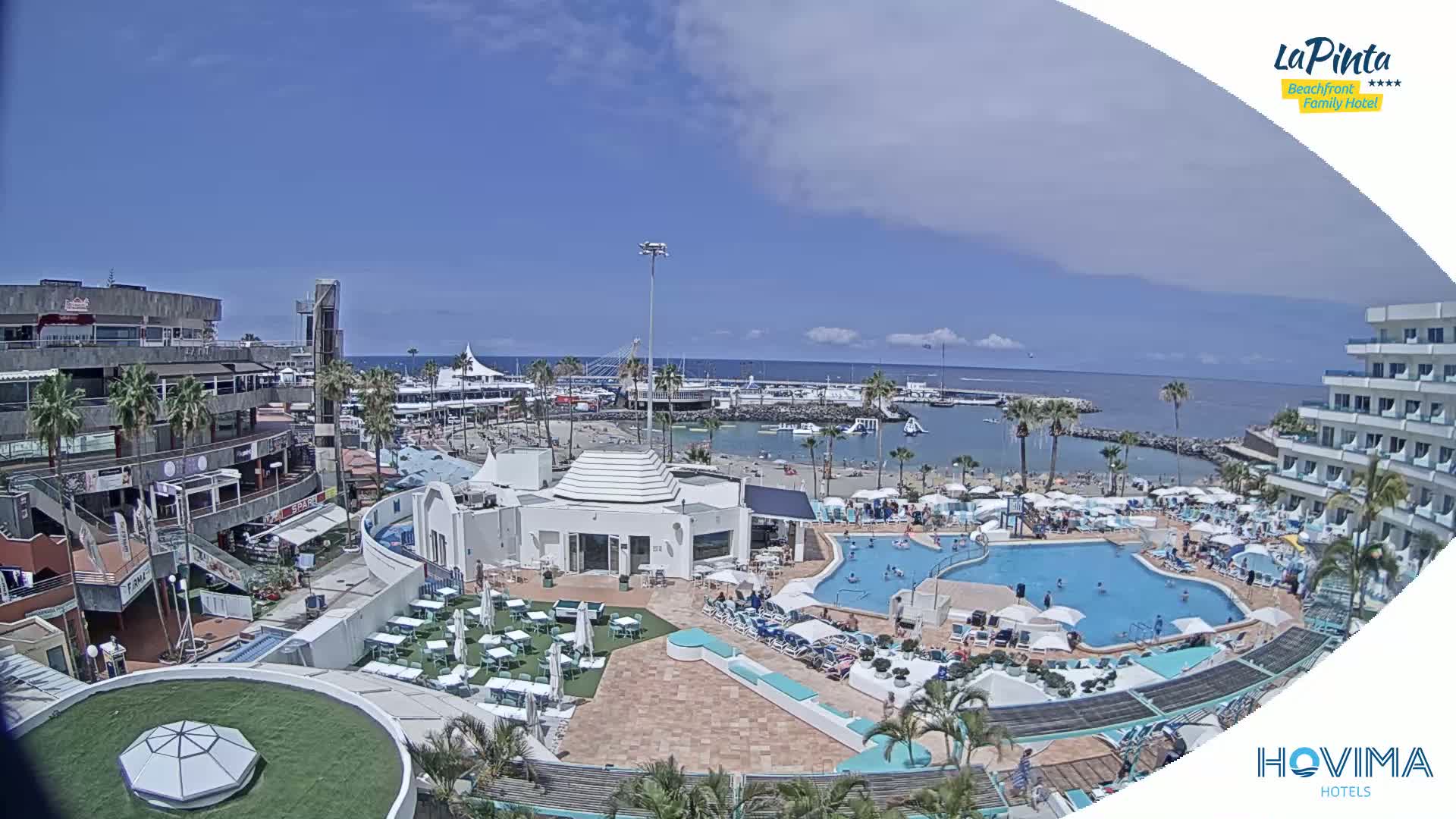 A resort complex with a swimming pool and beach access overlooks a marina on a sunny day.