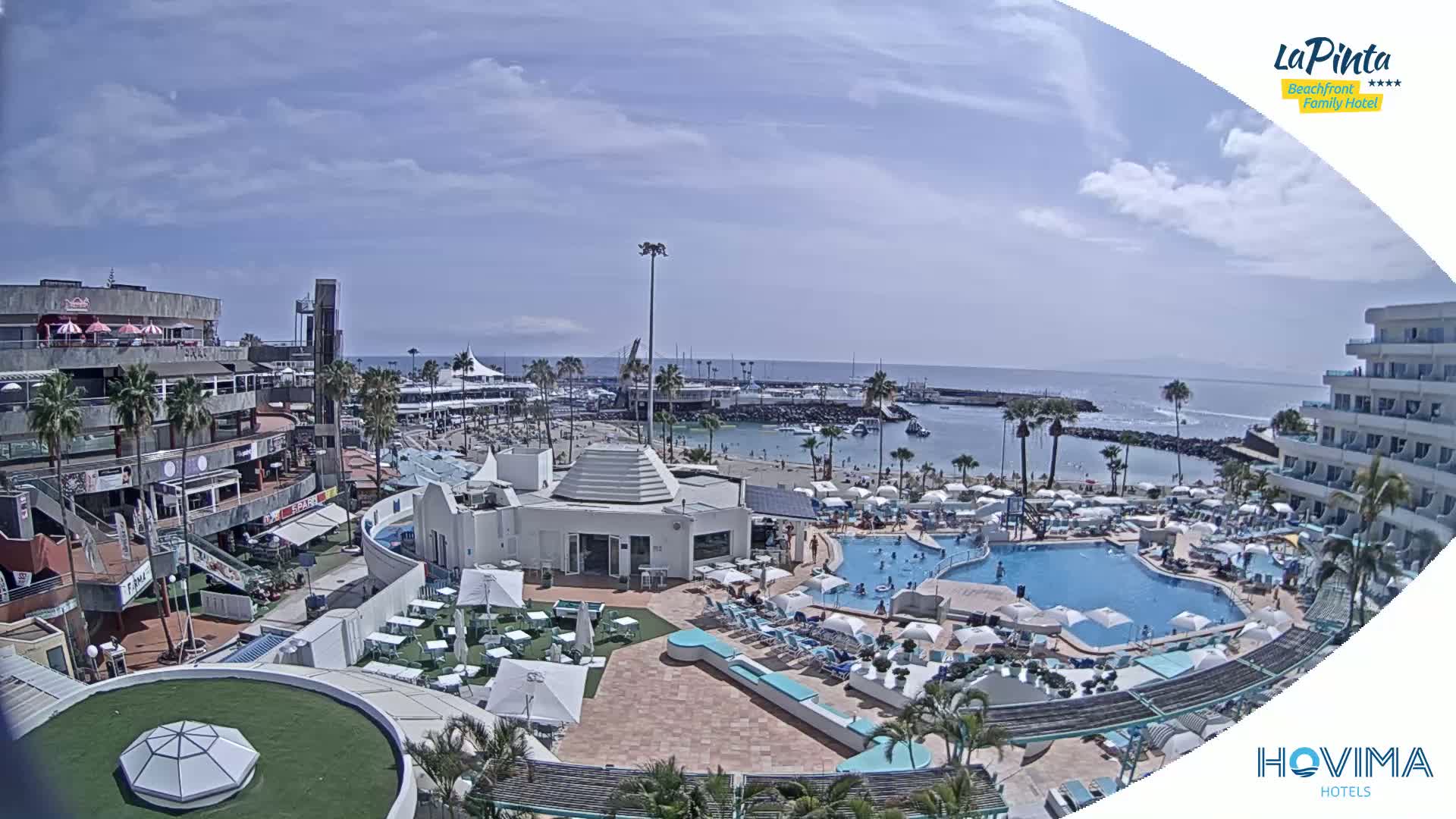 An aerial view of a resort with a swimming pool and beach, under a partly cloudy sky.