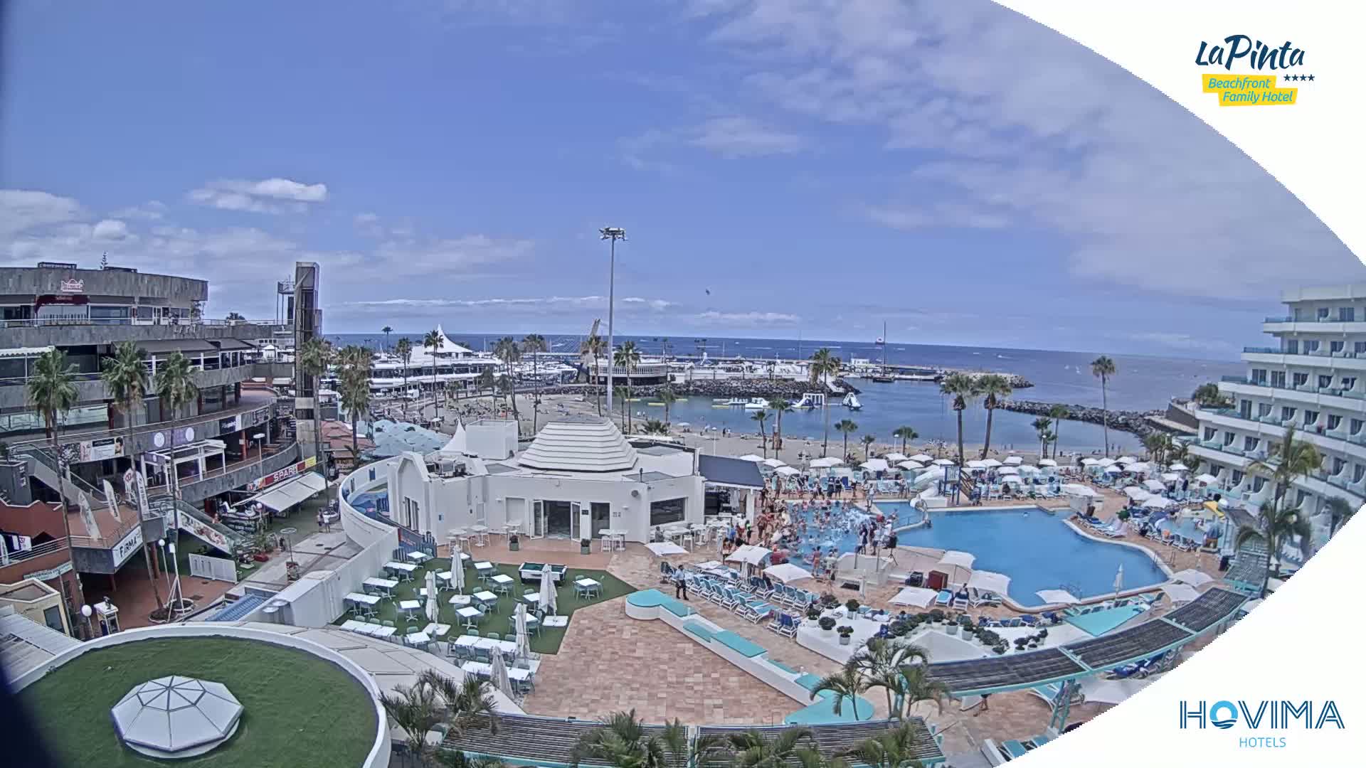 A sunny resort scene shows a swimming pool, beach, harbor, and buildings, with many people enjoying the outdoors.