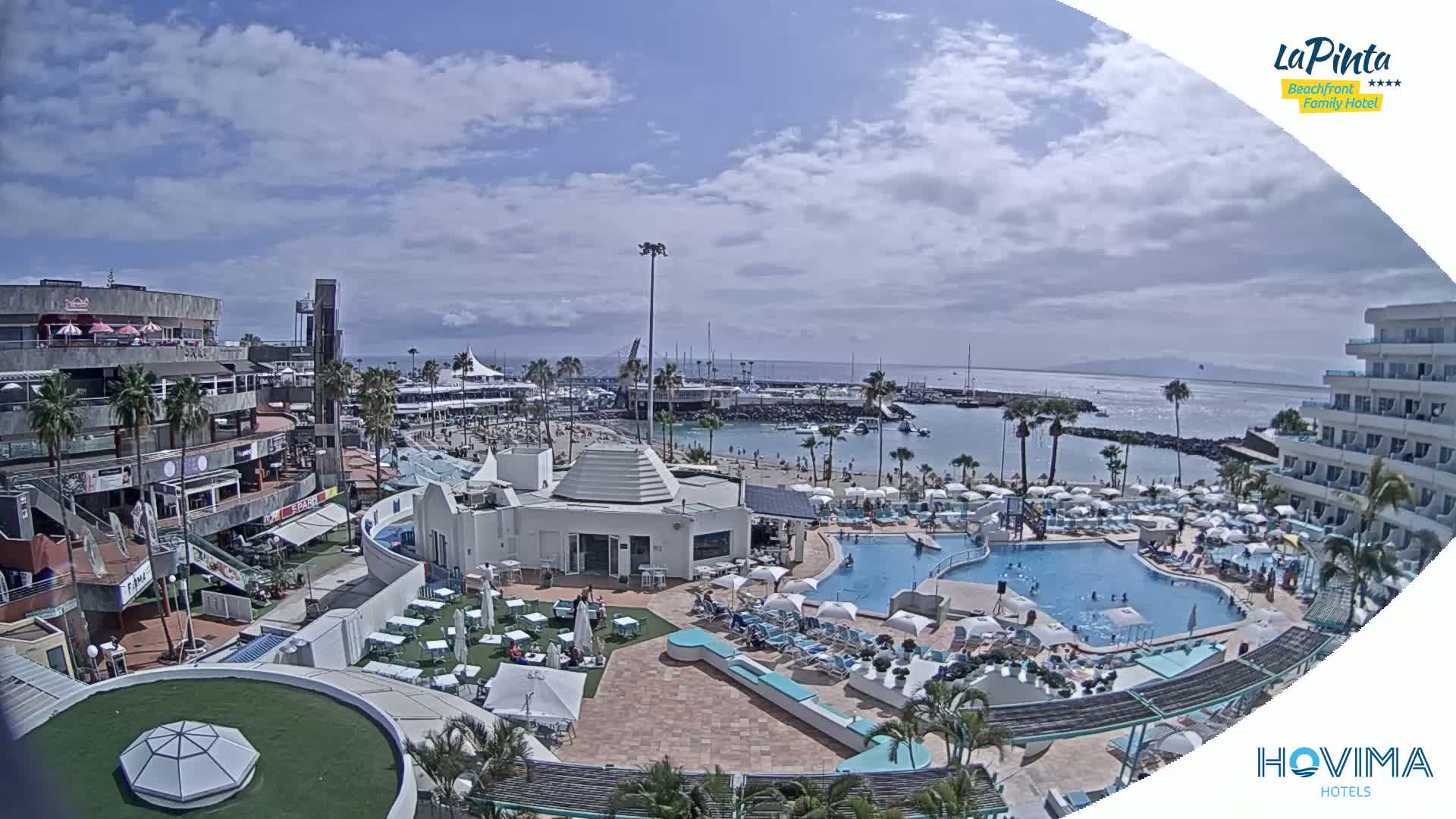 A sunny day overlooking a resort area with a swimming pool, beach, and marina, showing partly cloudy skies.