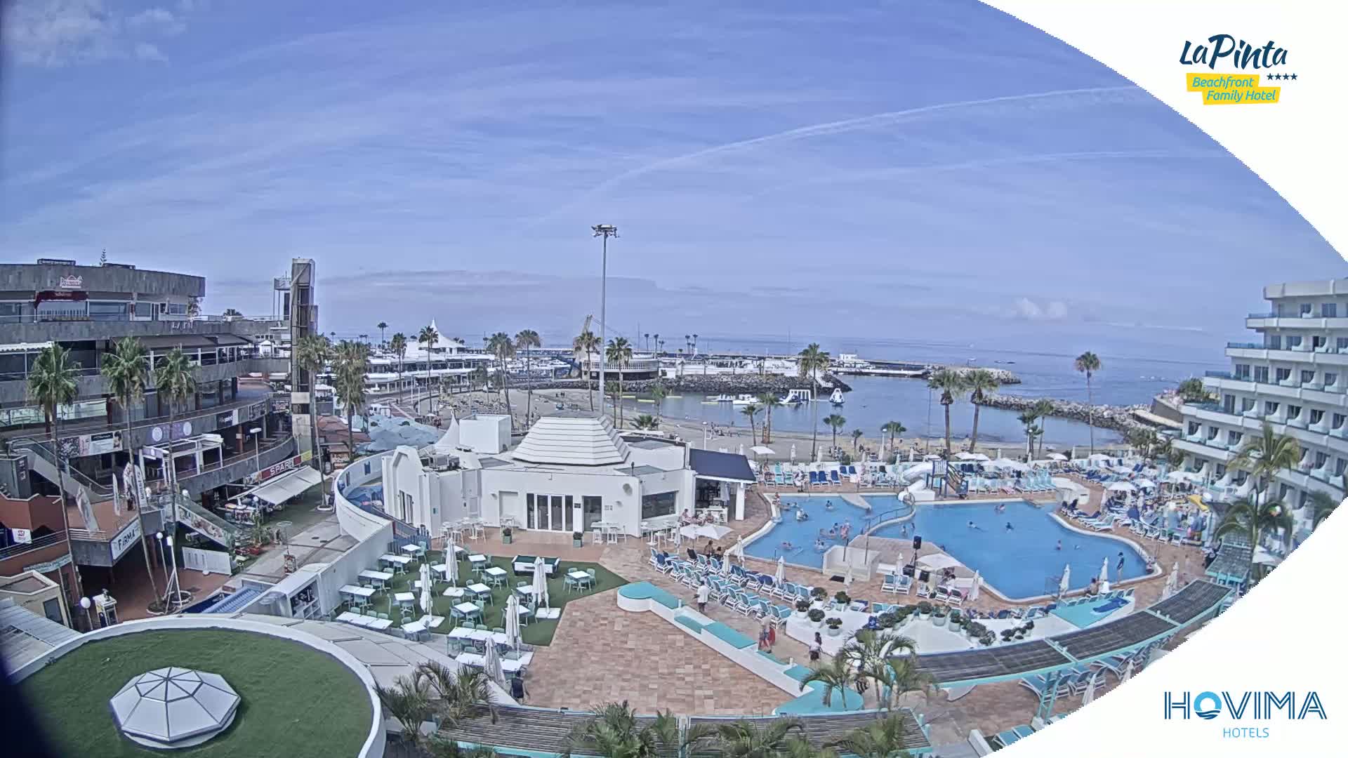 A sunny day overlooks a resort with multiple pools, a beach, and a marina; buildings and shops are visible in the background.