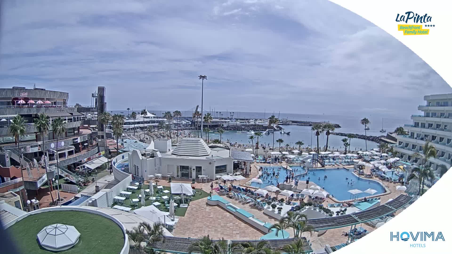 A resort area with a swimming pool and beach, under a partly cloudy sky, overlooks a calm harbor with boats.