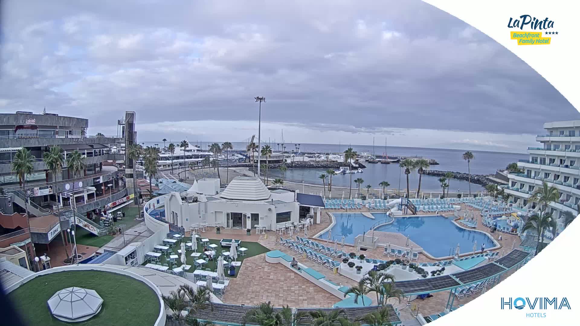 An overcast day reveals a resort with multiple pools and a small beach, overlooking a calm marina and ocean.
