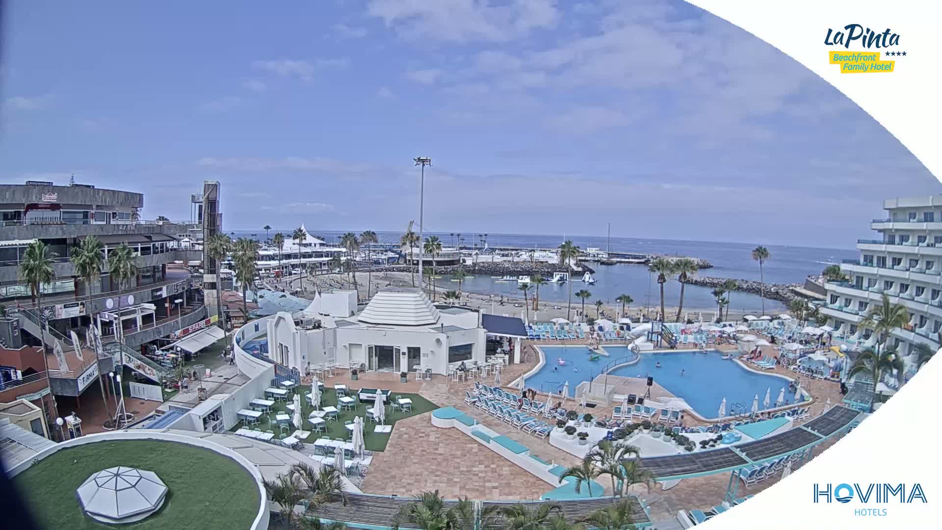 A resort pool area with a small beach and calm ocean in the background under a mostly sunny sky.