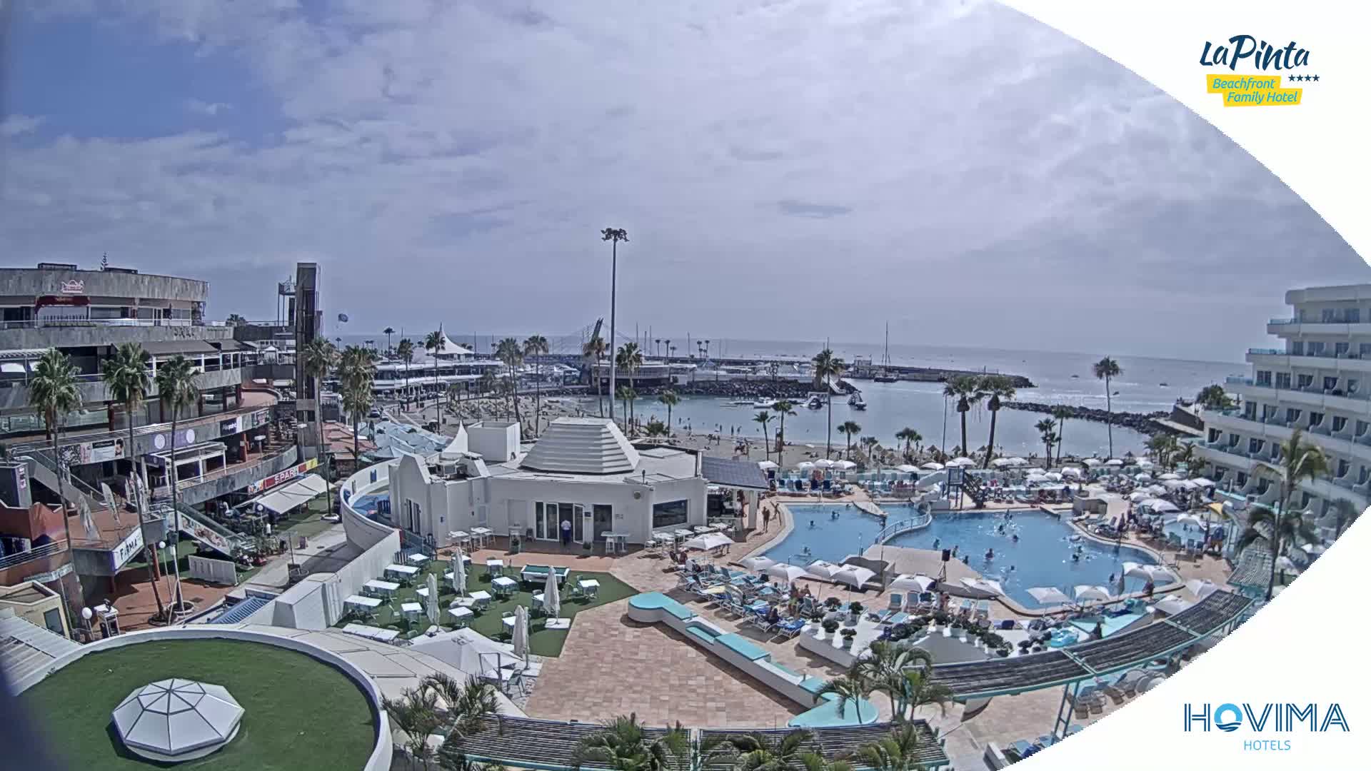 An aerial view shows a resort with multiple pools and a beach overlooking a calm ocean under a partly cloudy sky.