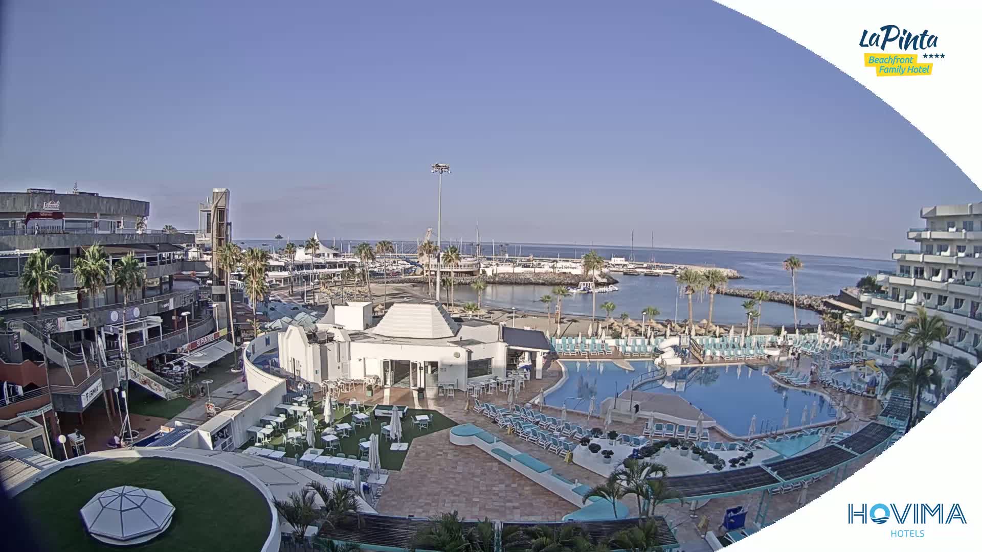 A resort with a swimming pool overlooks a marina and calm ocean under a clear blue sky.