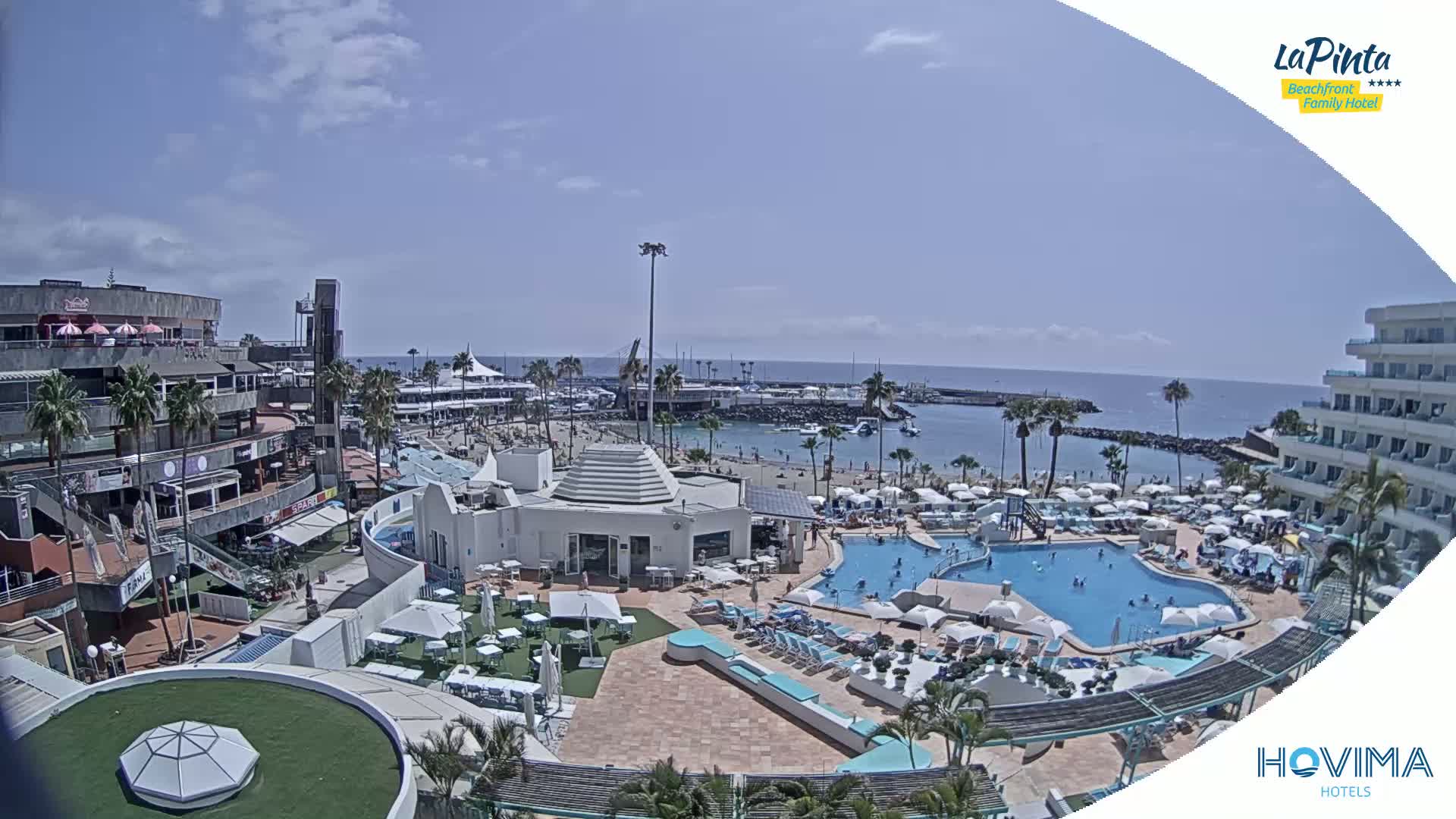 A resort with multiple pools and a beach area is visible, along with shops and buildings nearby, all under a sunny sky.