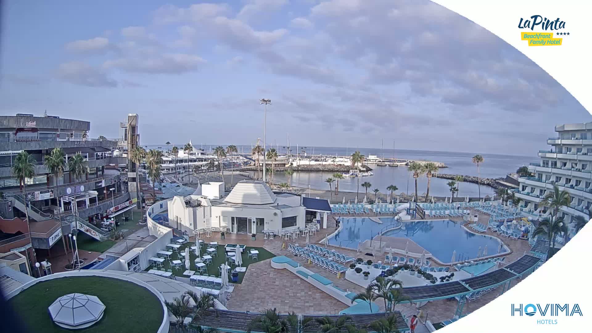 A resort pool area with a marina and ocean view under a partly cloudy sky.