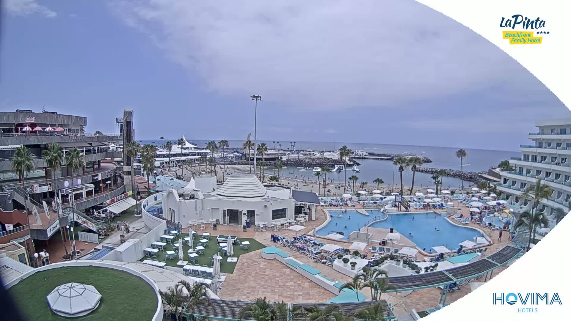 An aerial view of a resort area on a partly cloudy day shows a swimming pool, beach, and buildings surrounding a calm harbor.