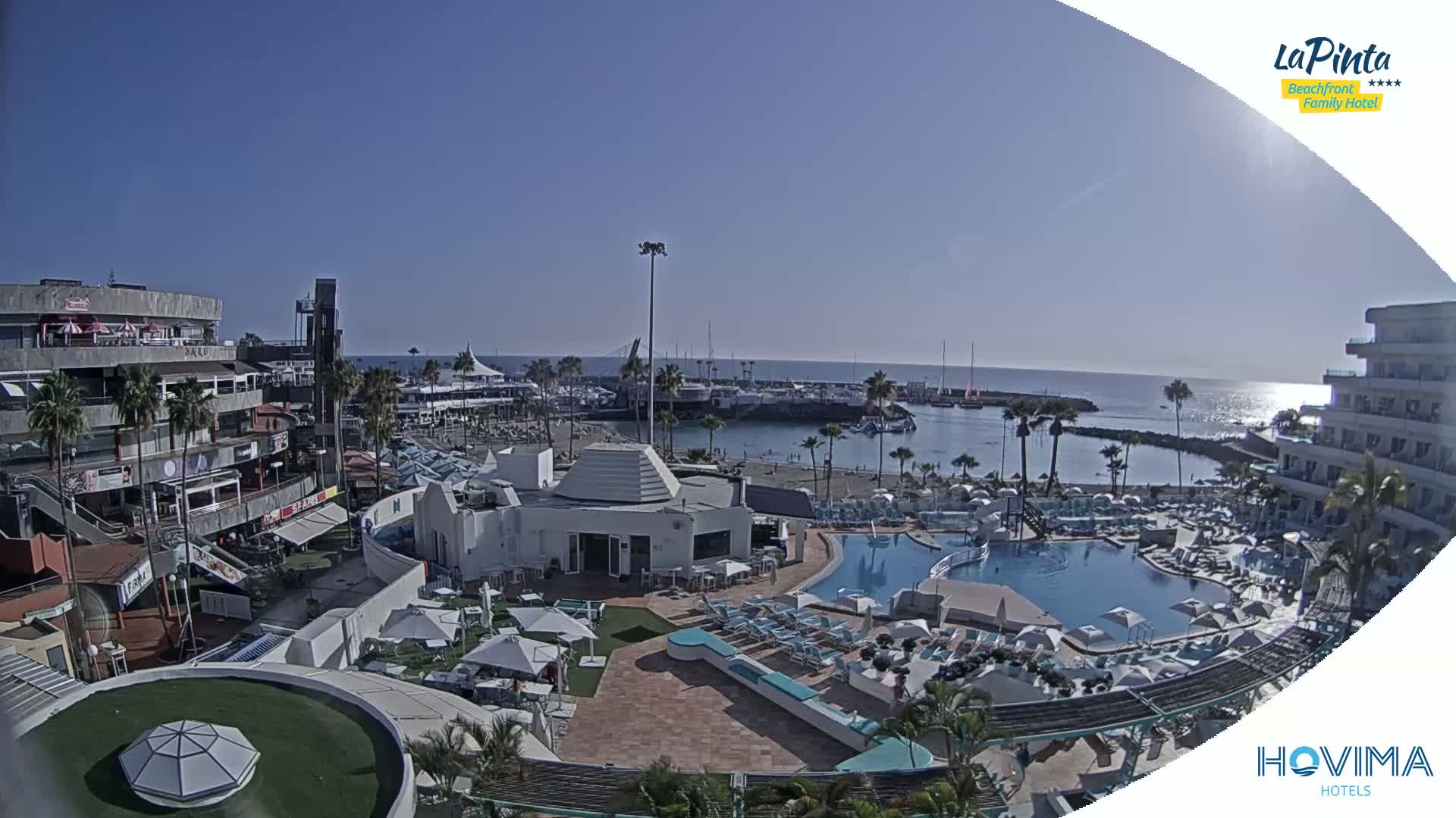 An aerial view shows a resort pool area next to a marina and beach on a sunny day.