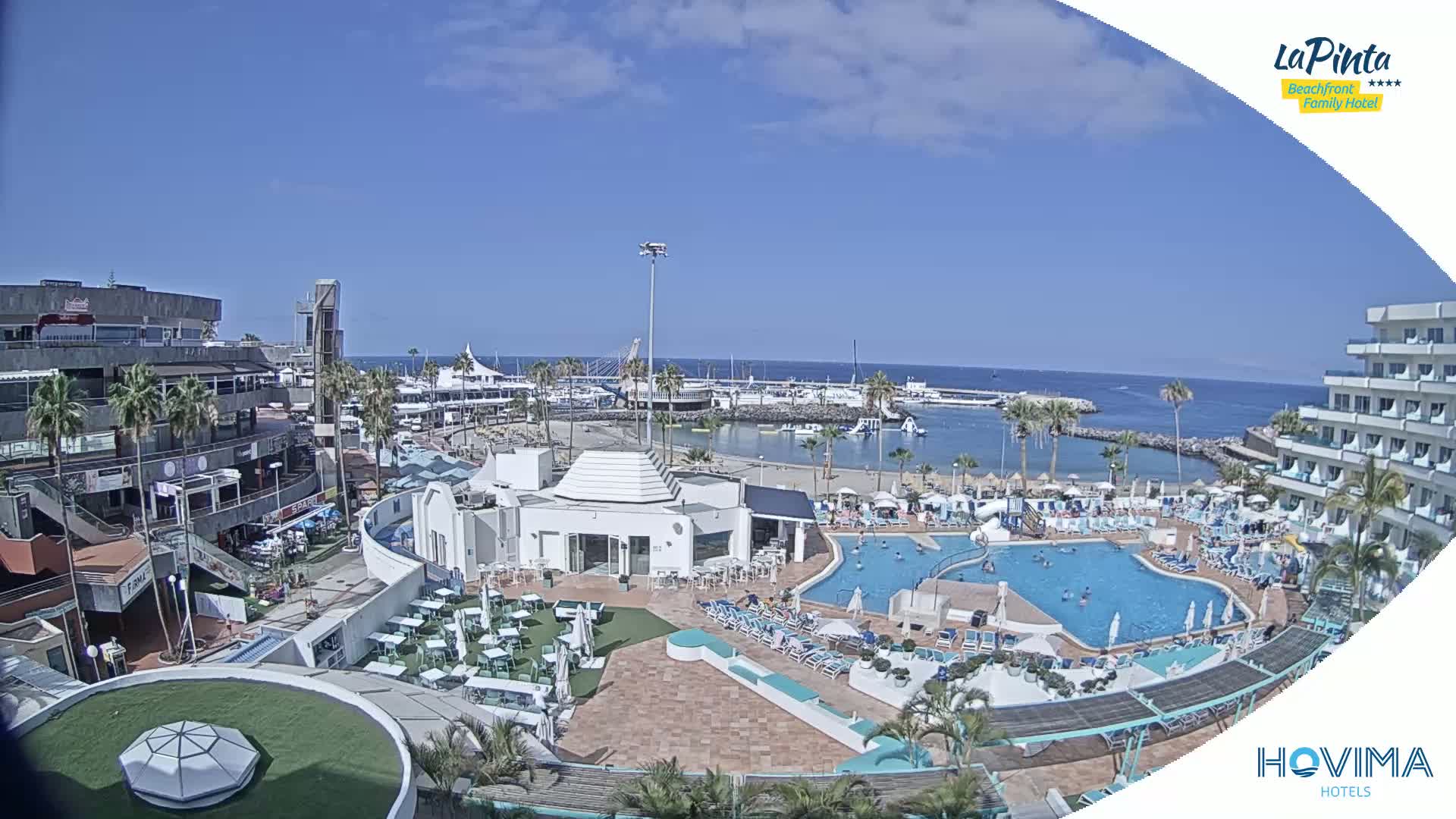 An aerial view of a resort complex on a sunny day, showing a swimming pool, beach area, marina, and buildings, all overlooking a calm ocean.