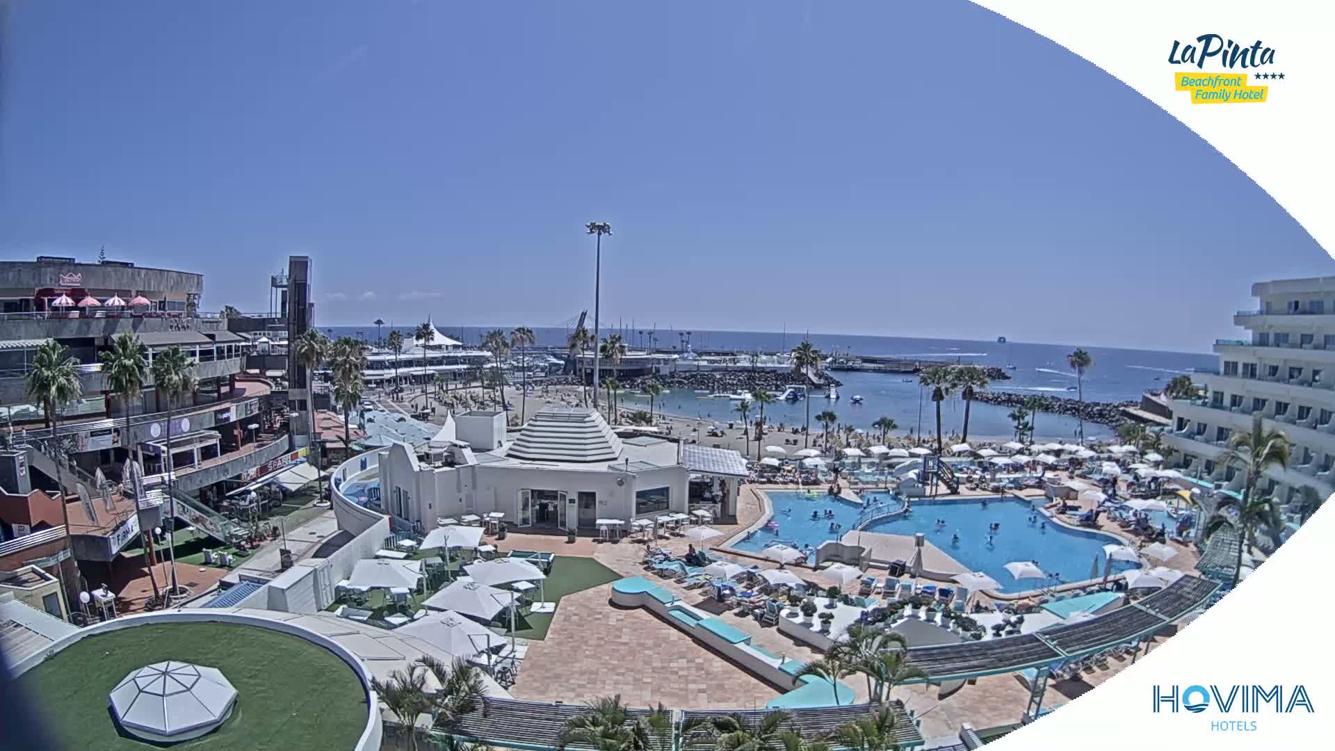 An aerial view of a resort on a sunny day shows a beach, several pools, and a collection of buildings near a calm ocean.