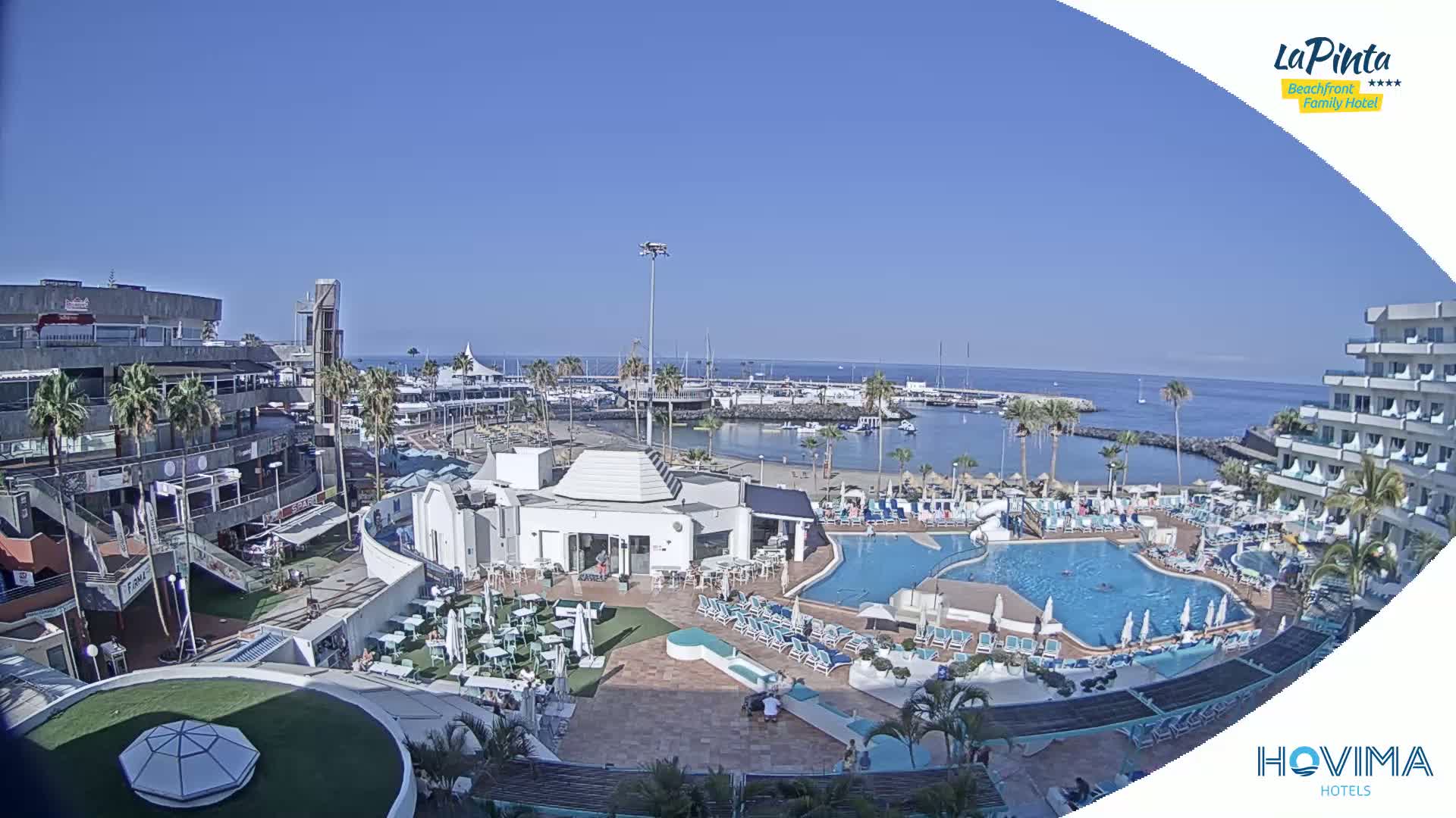 A sunny outdoor scene shows a resort area with a swimming pool, beach, marina, and shops.