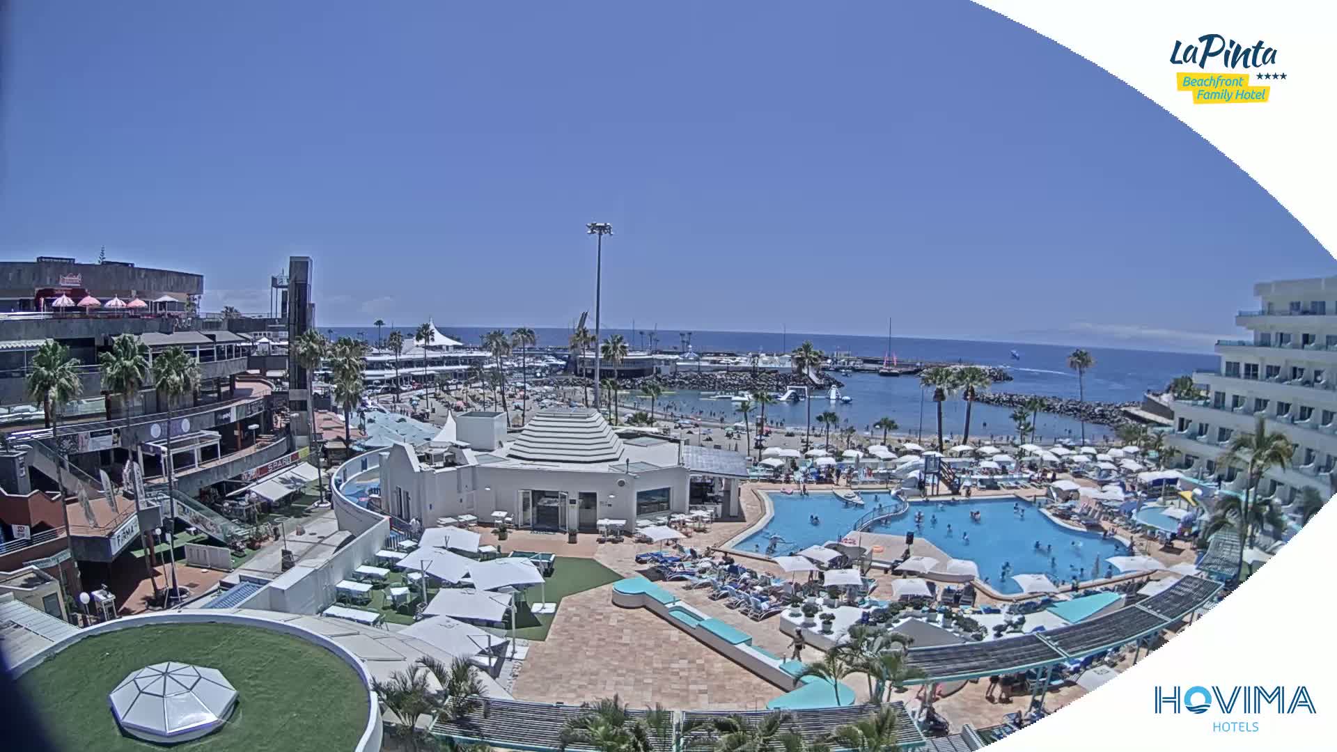 A sunny day at a resort overlooking a calm ocean, featuring a pool area, beach, and various buildings.