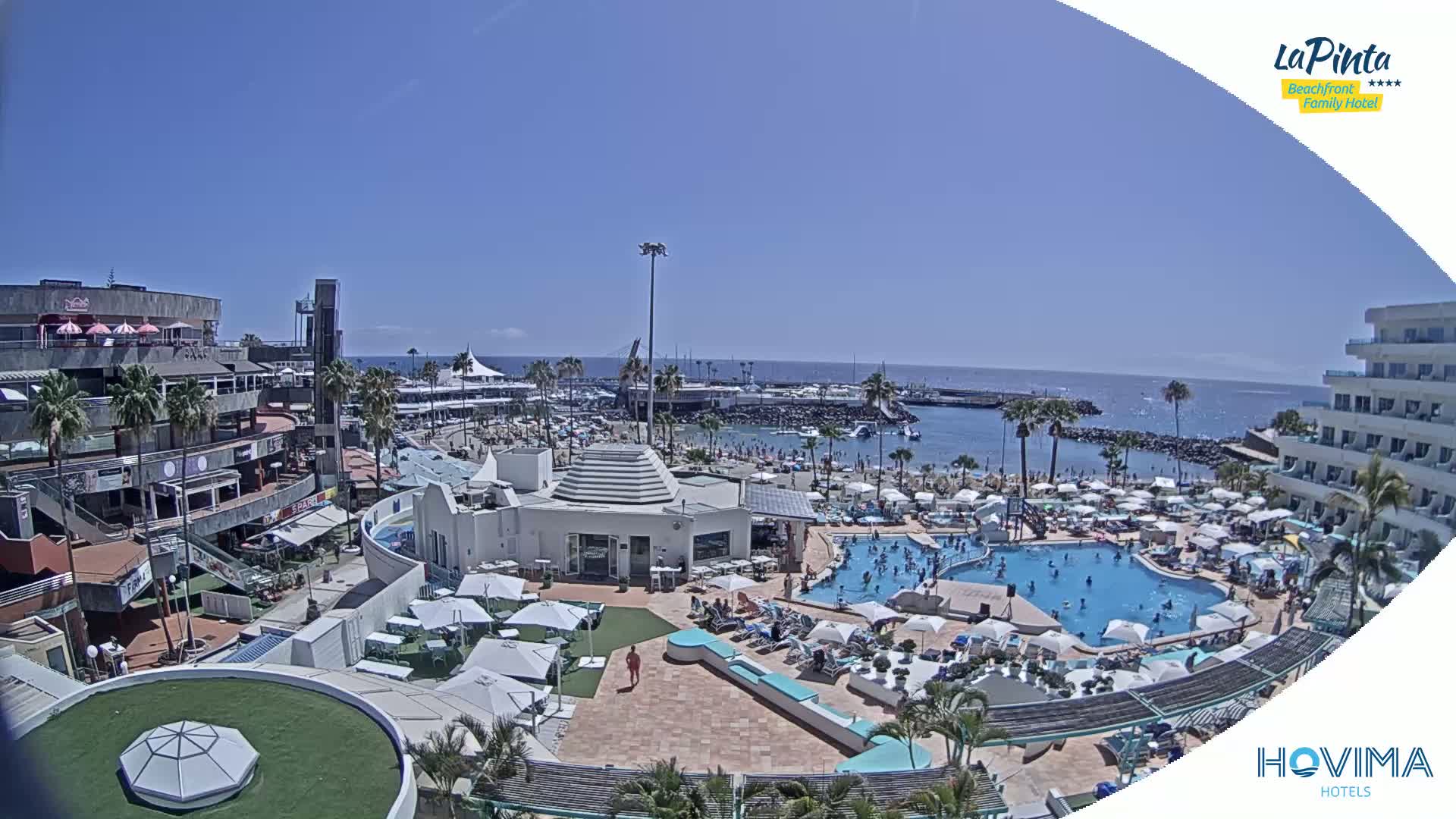 A sunny beach resort with a large pool and many people enjoying the water, surrounded by buildings and a calm ocean.