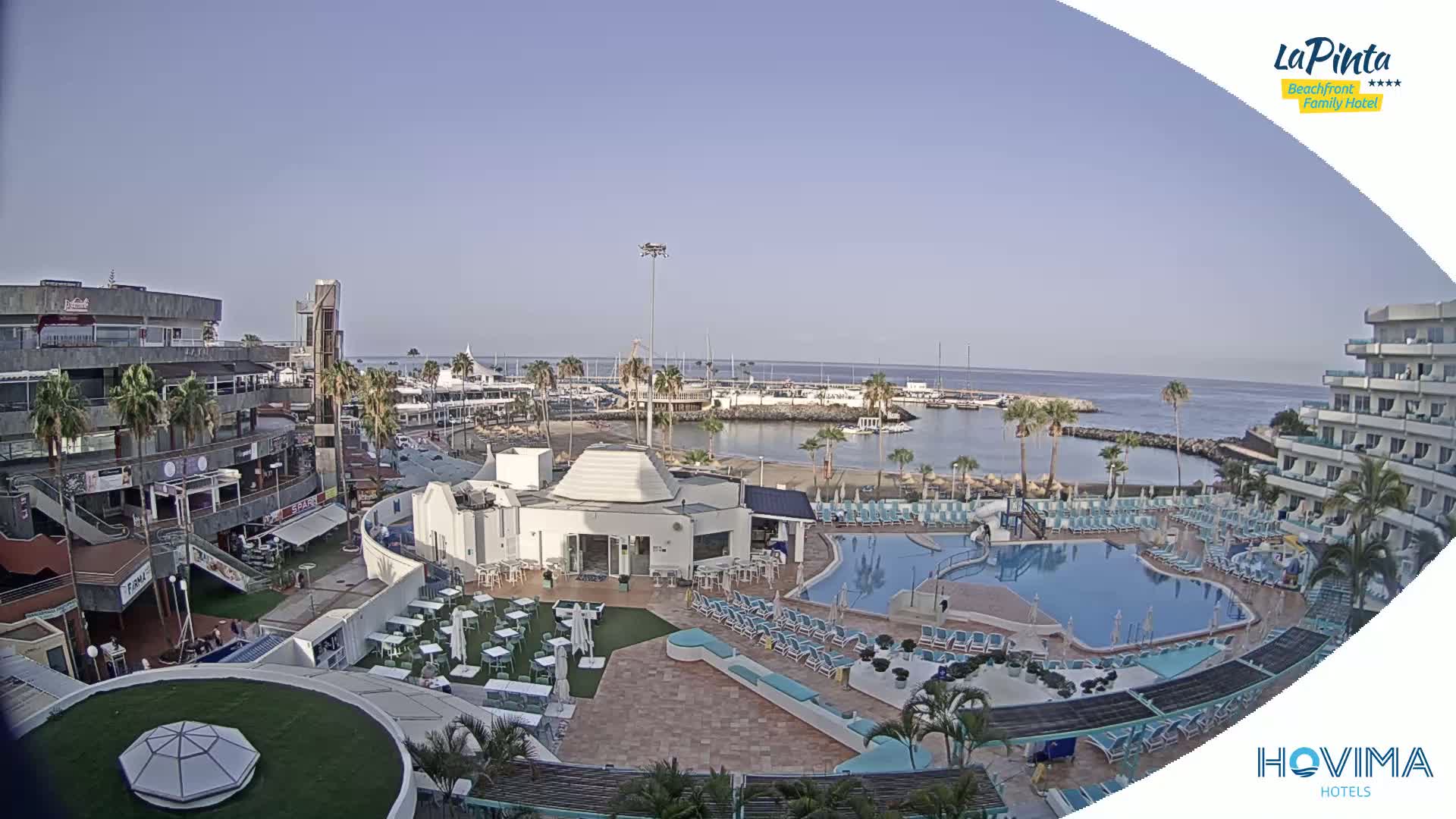 A resort complex with a swimming pool overlooks a calm harbor filled with boats under a clear sky.